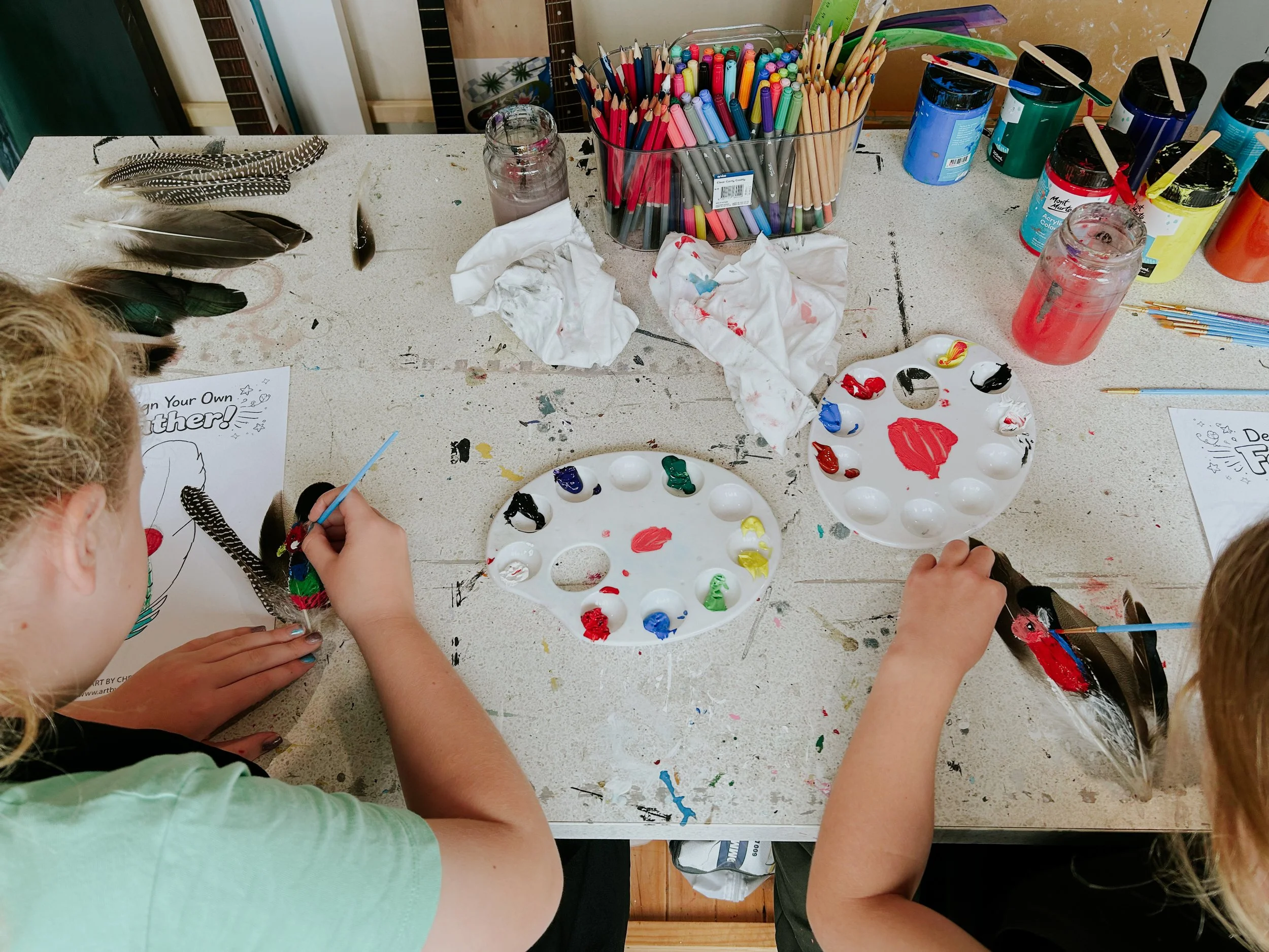 Children engaging in an art project at a crafting table with feathers, paints, colored pencils, and drawing materials.
