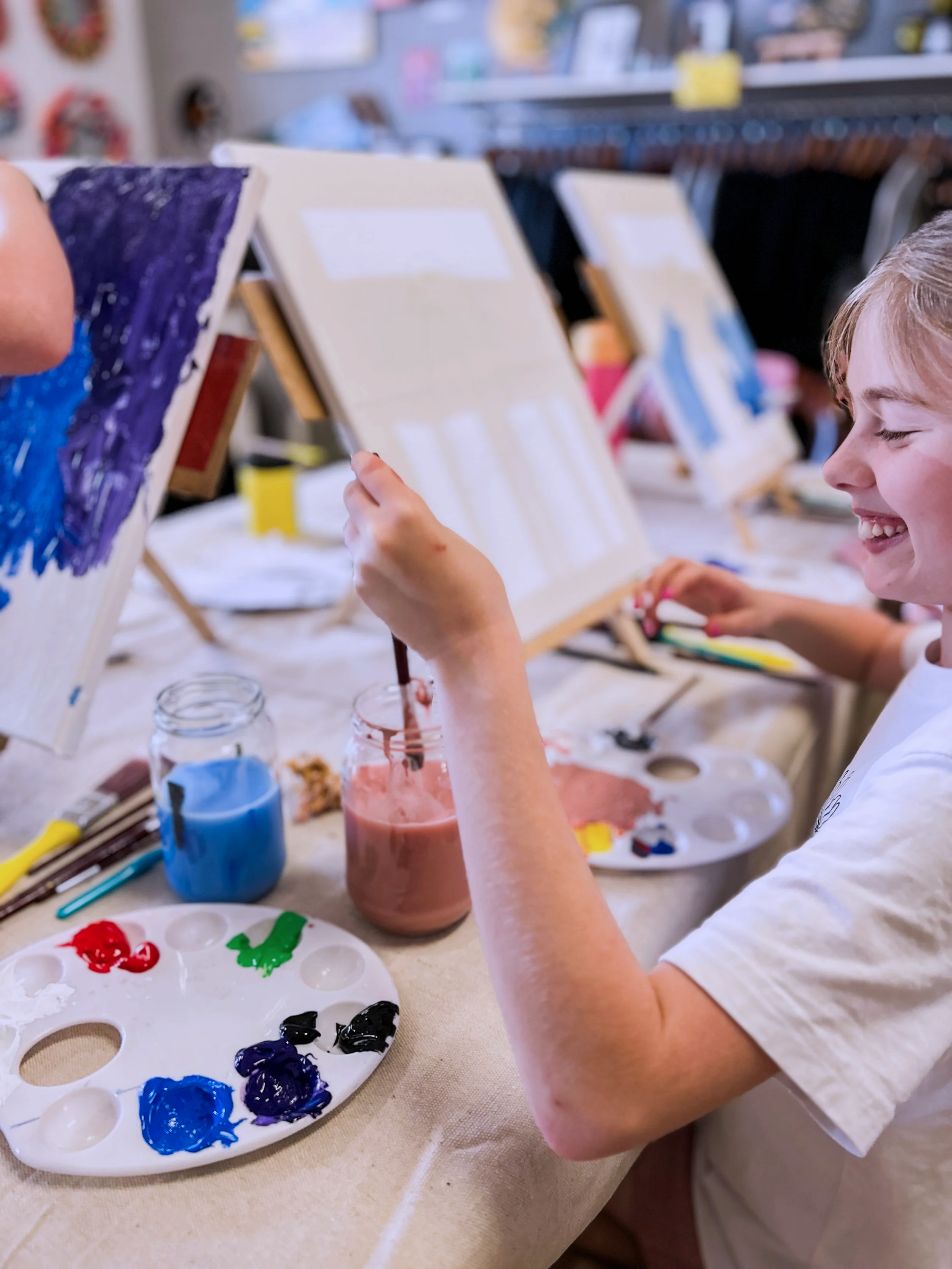A young girl with blonde hair is painting on a canvas with pink paint, smiling. The canvas is on an easel, and there are paint palettes, jars of colored paints, and brushes on the table in front of her. Another canvas with blue and purple paint is visible on the left.