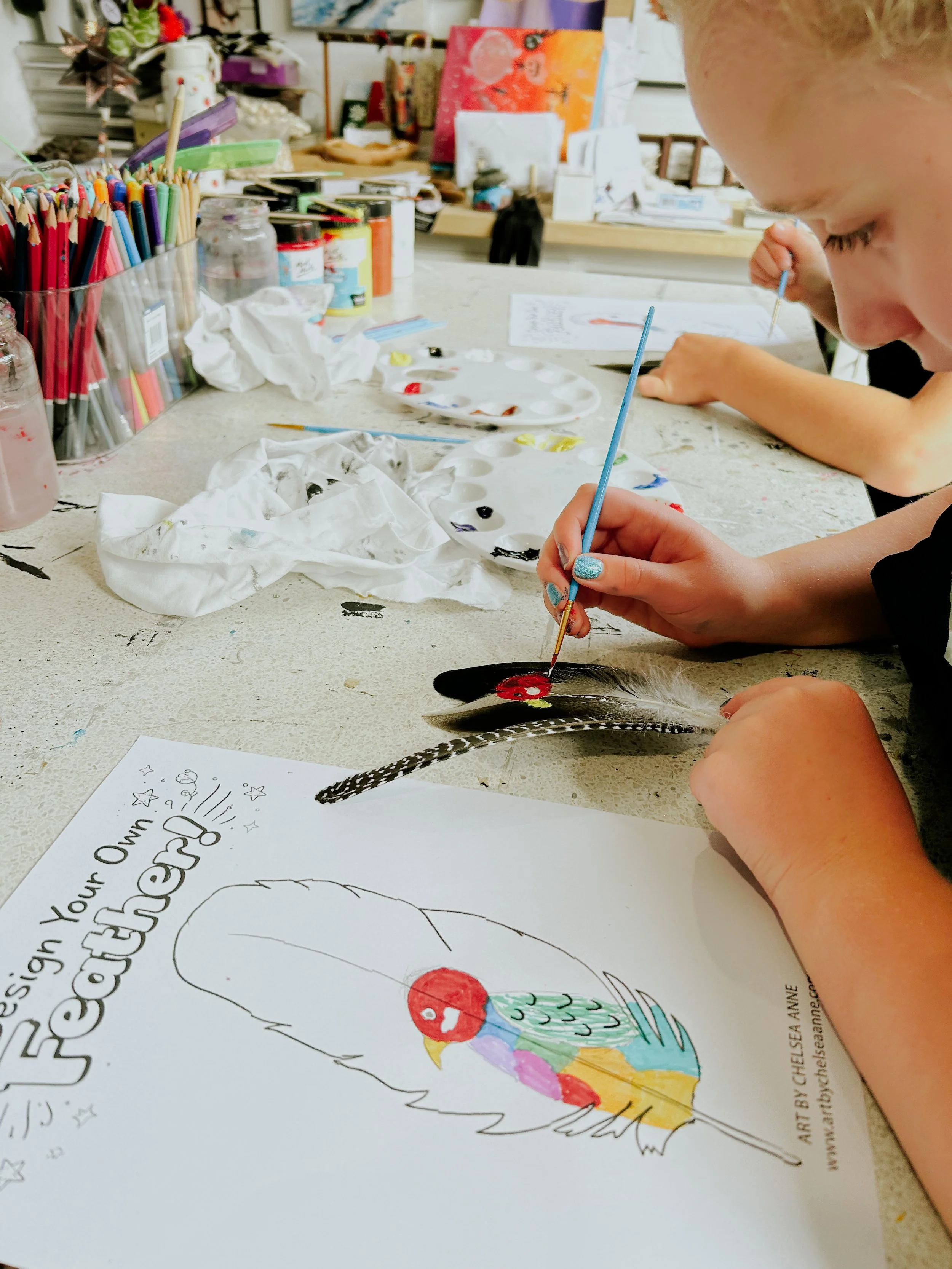 A young girl is painting a colorful bird on a sheet of paper in an art studio. The table is cluttered with paint supplies, and in the background, there are canvases and art materials.