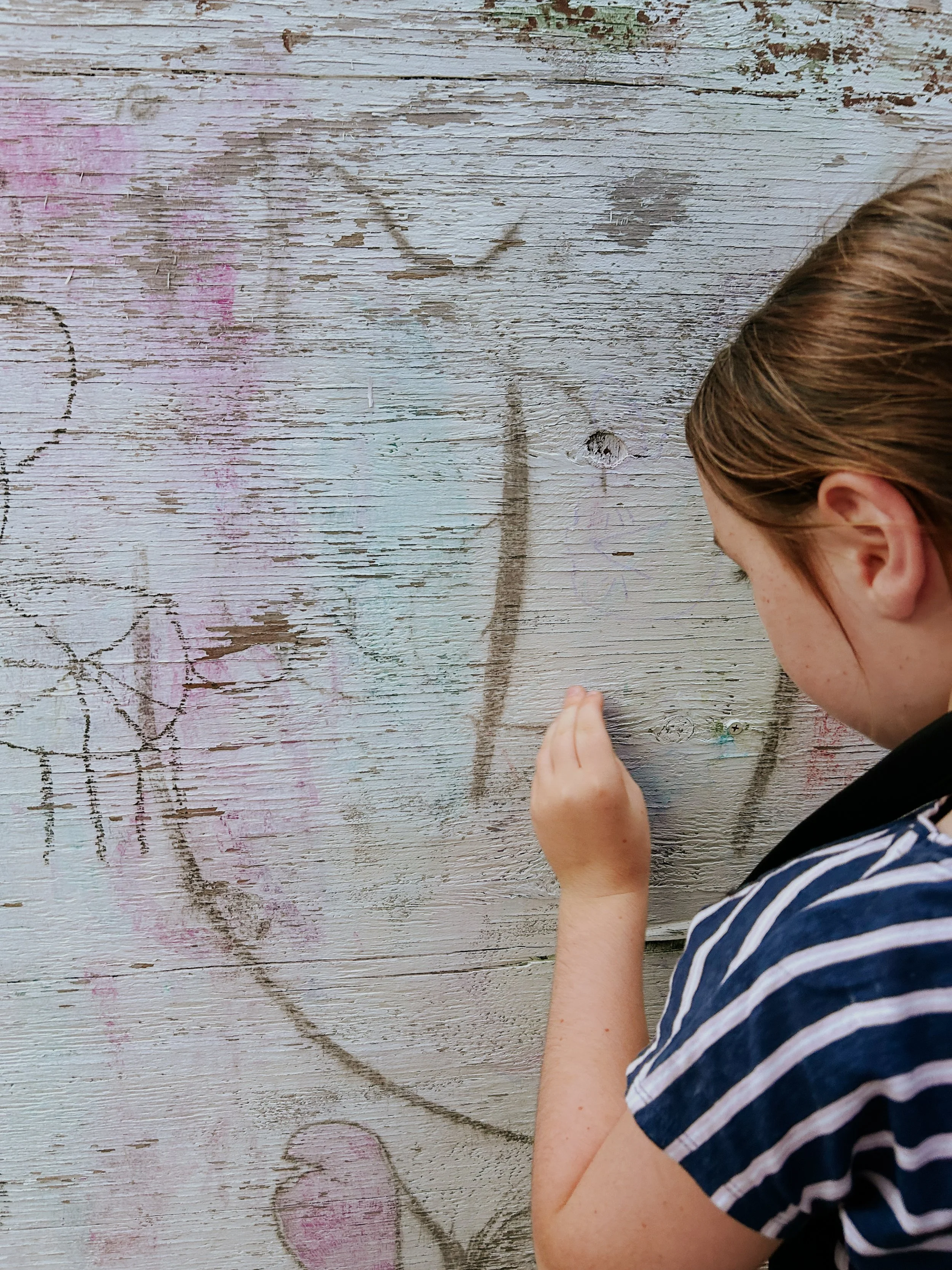 A young girl with reddish-brown hair wearing a blue and white striped shirt, working on a drawing on a weathered wooden wall.