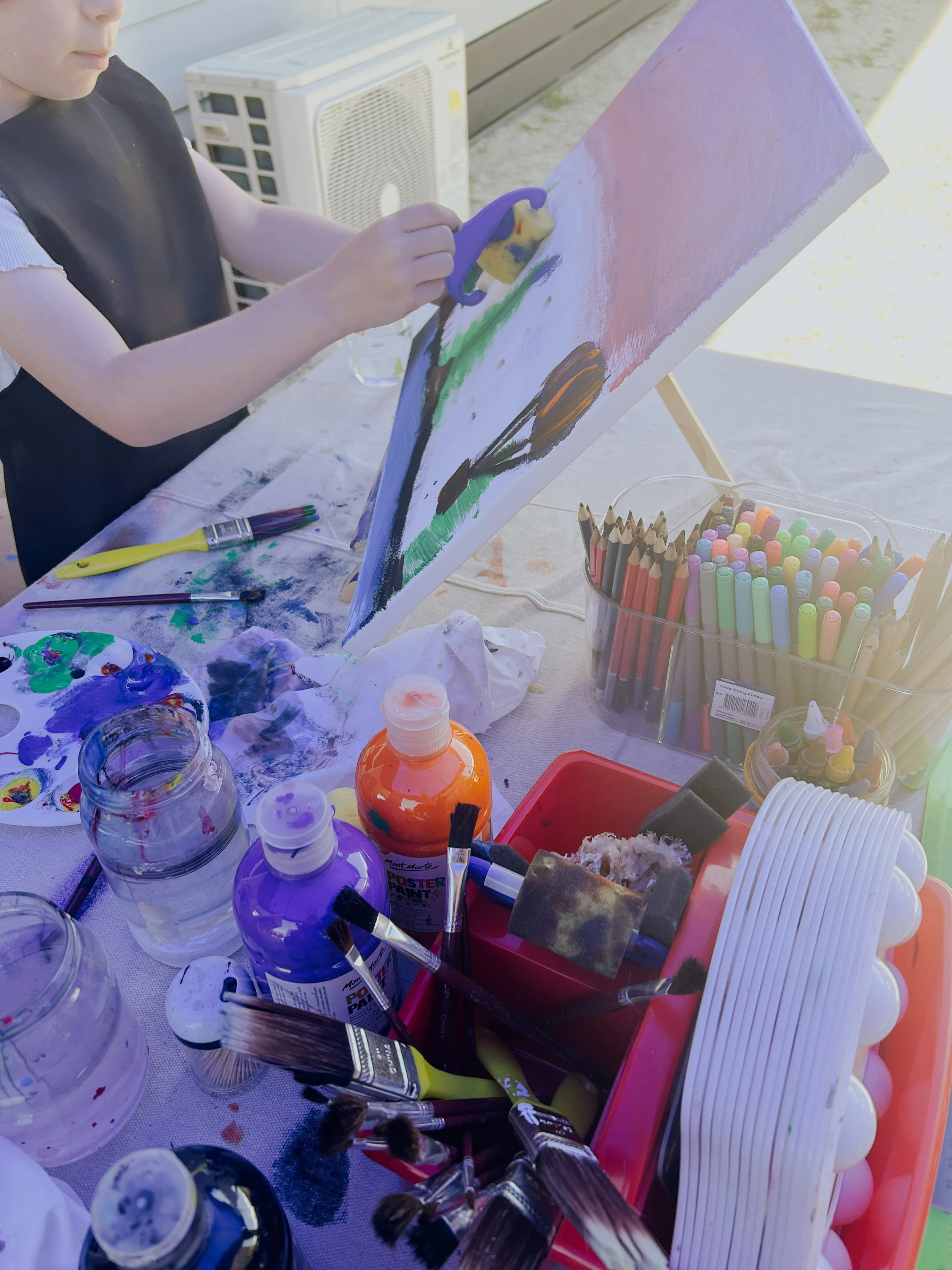 A child painting on a canvas outdoors with various art supplies on a table, including paint bottles, brushes, colored markers, and a palette.