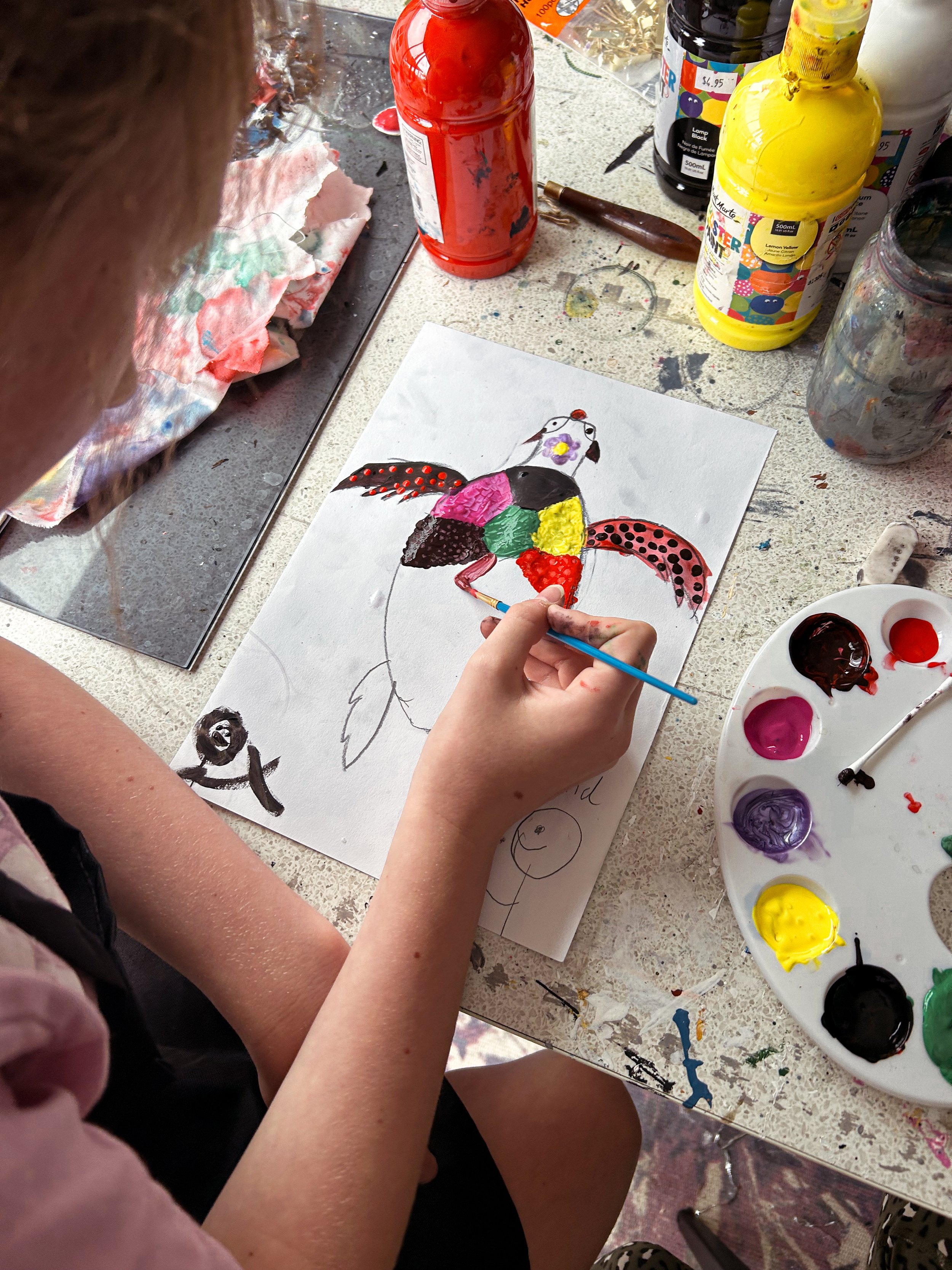 Child painting a colorful, abstract parrot on paper with various art supplies on the table.