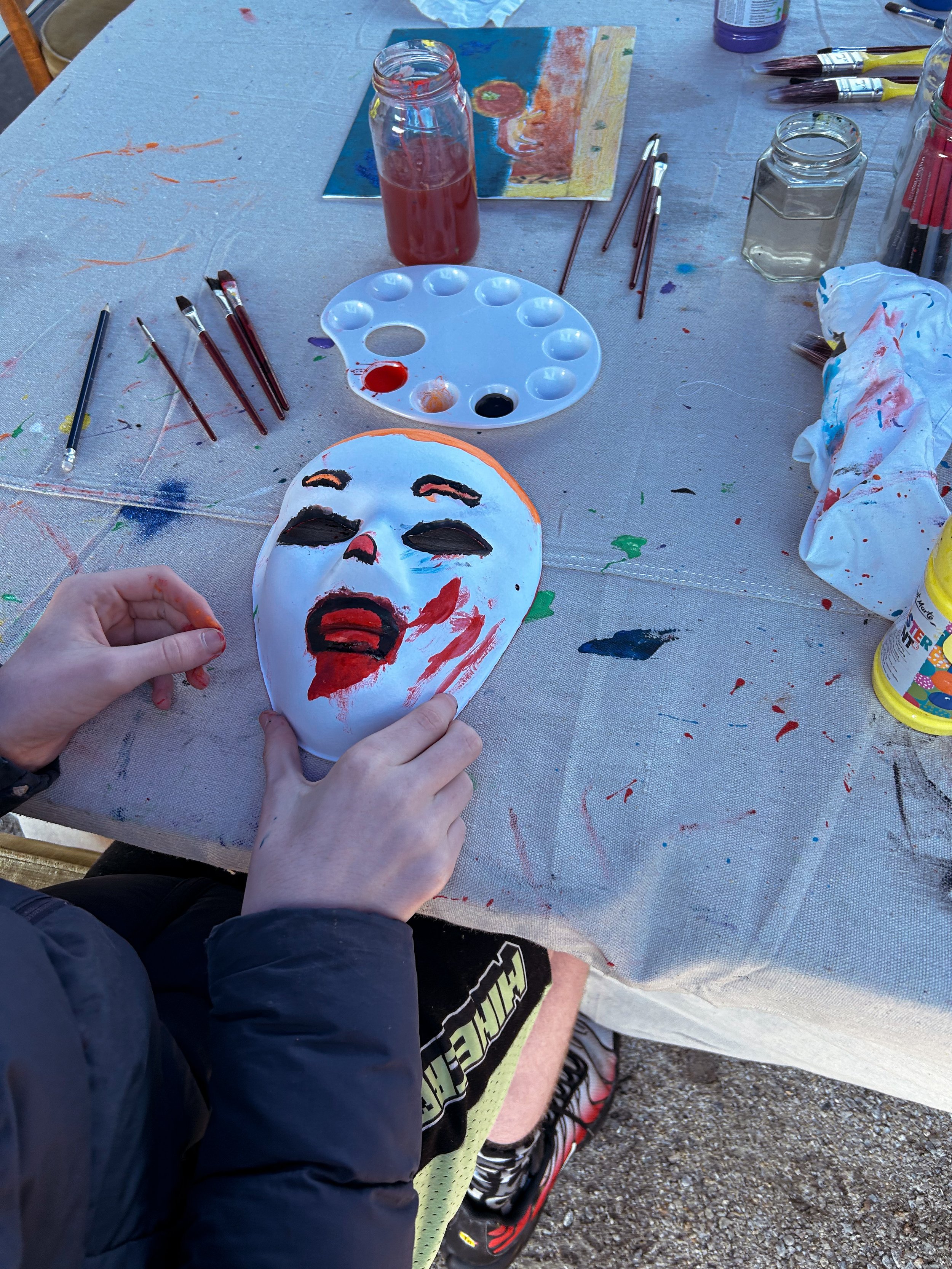 A person painting a spooky clown mask with black eyes, red mouth, and red streaks on a white mask, surrounded by paint supplies and brushes on a table.