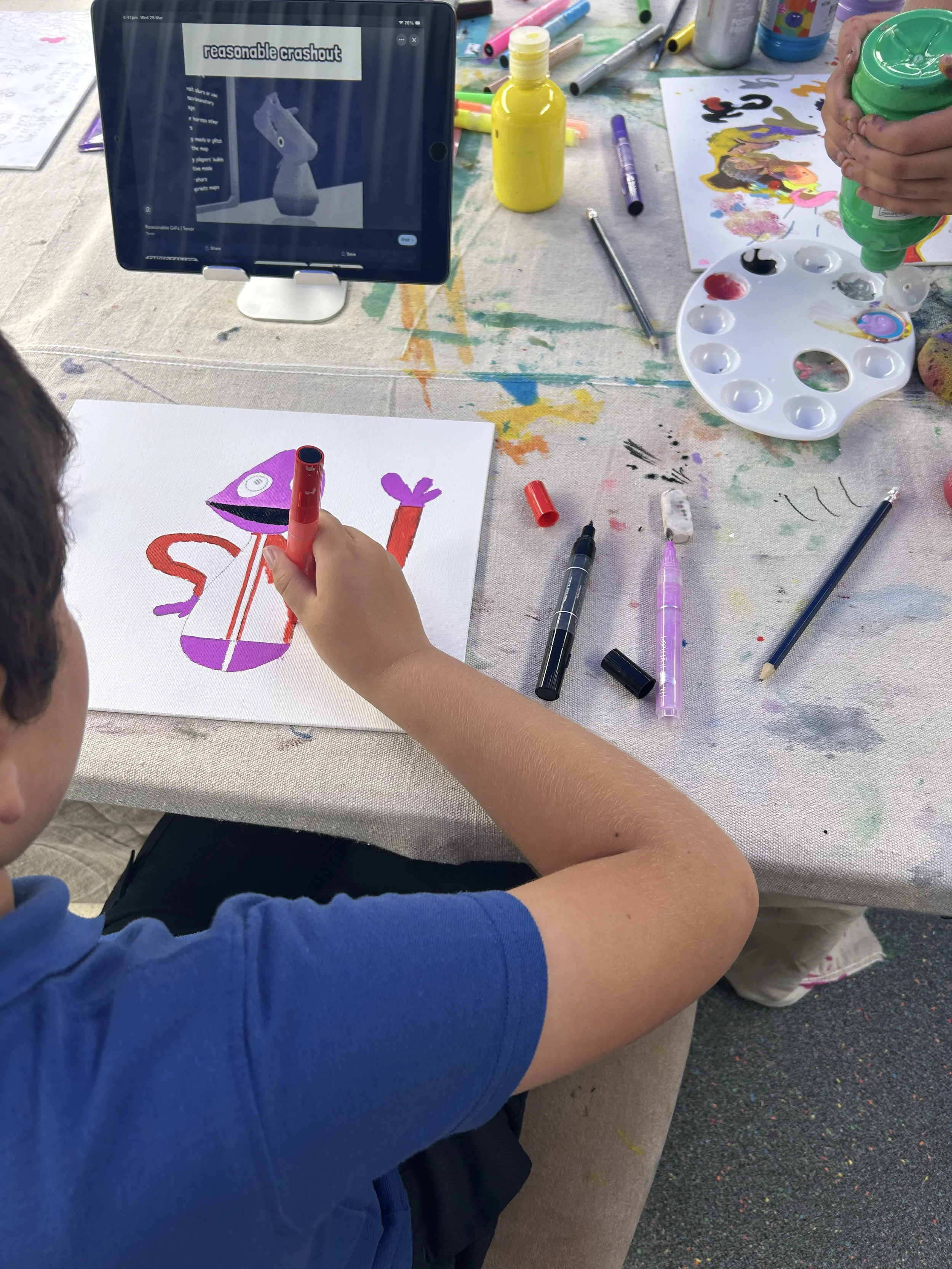 Child using a red marker to draw a colorful cartoon-style character on white paper, with art supplies and a tablet showing an art tutorial in the background.