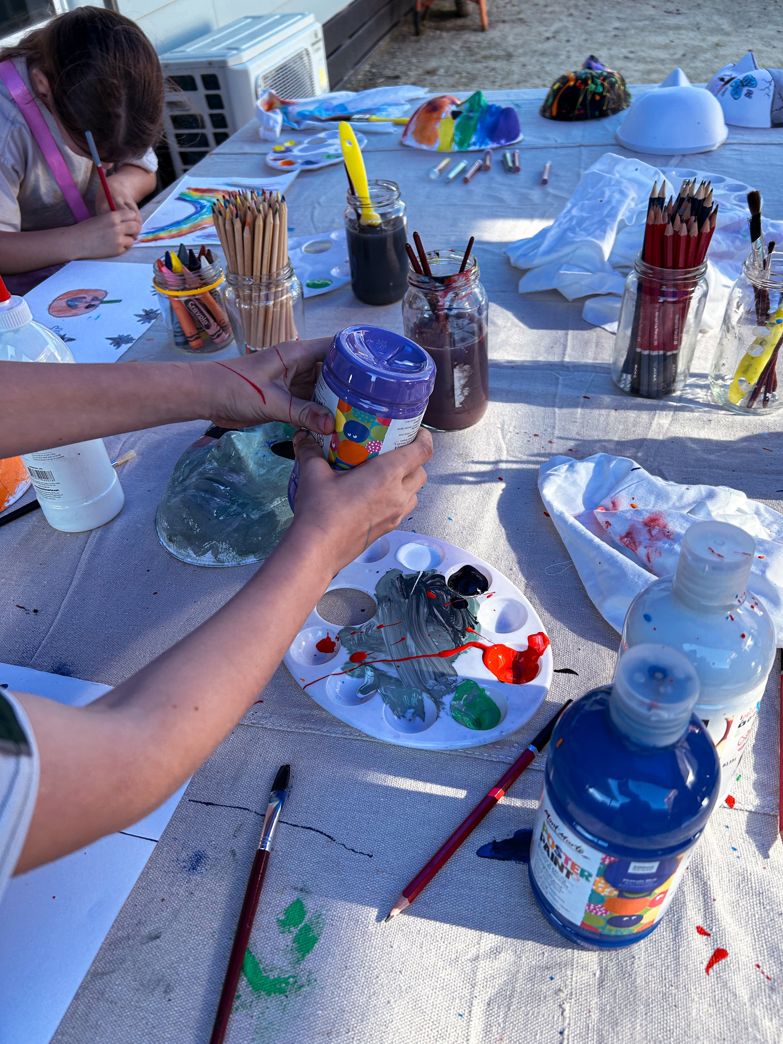 Children at an outdoor art station with paint jars, paintbrushes, and paper, creating colorful paintings.