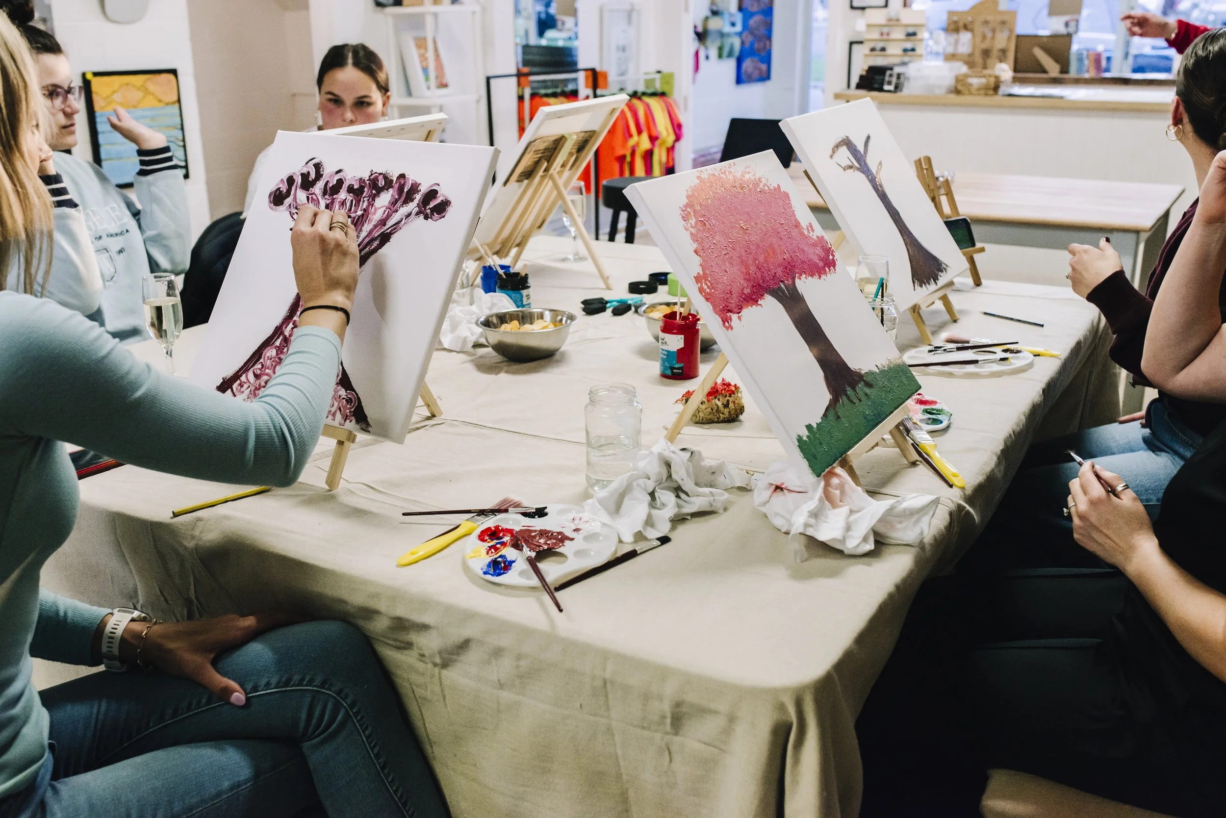 People participating in a painting class, working on individual canvases with various colorful landscape and tree paintings. Art supplies and paint palettes are on the table.