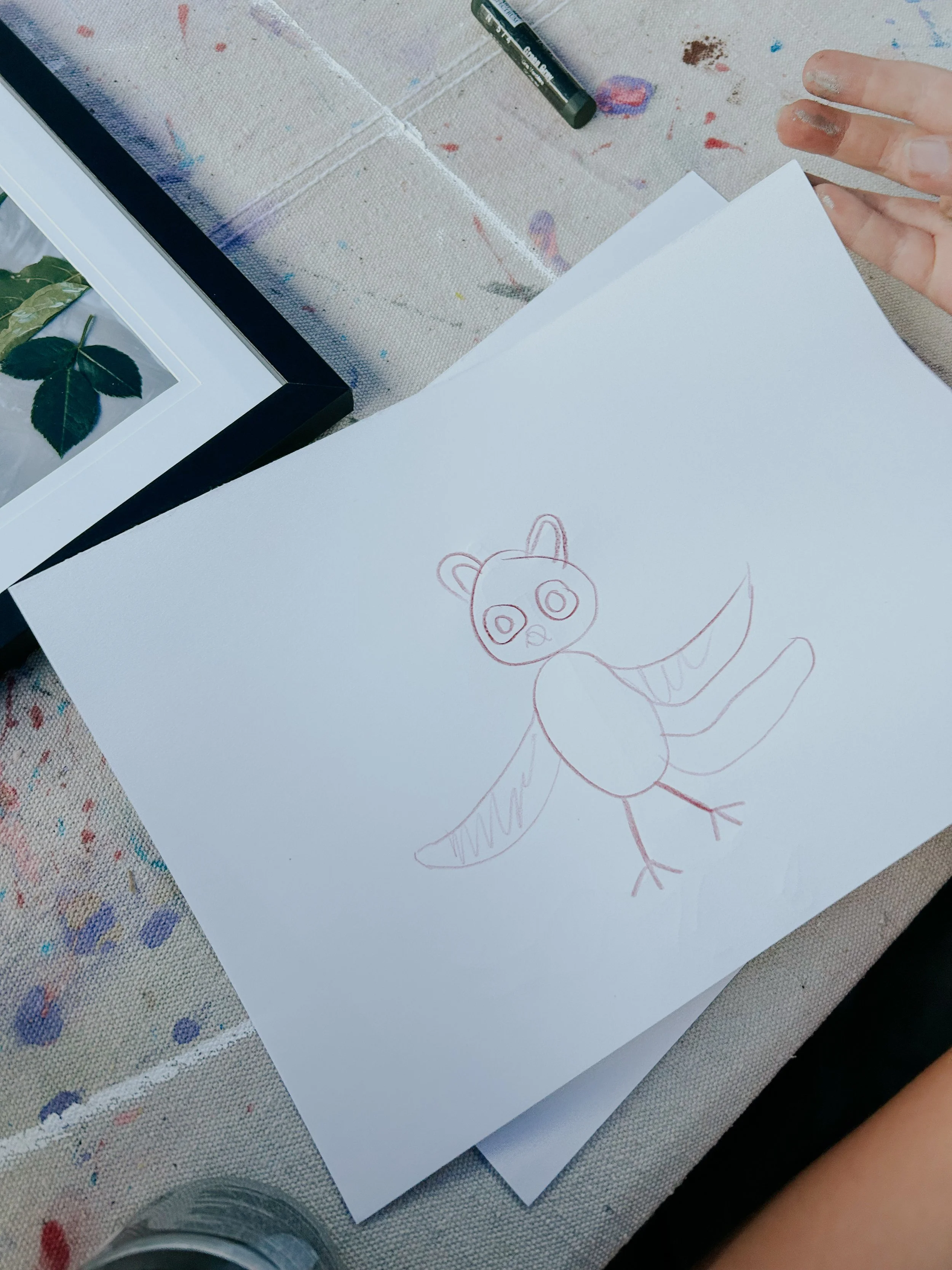 A hand with finger paint smudges reaching towards a piece of white paper with a drawing of an animal on it, on a colorful, splattered table.