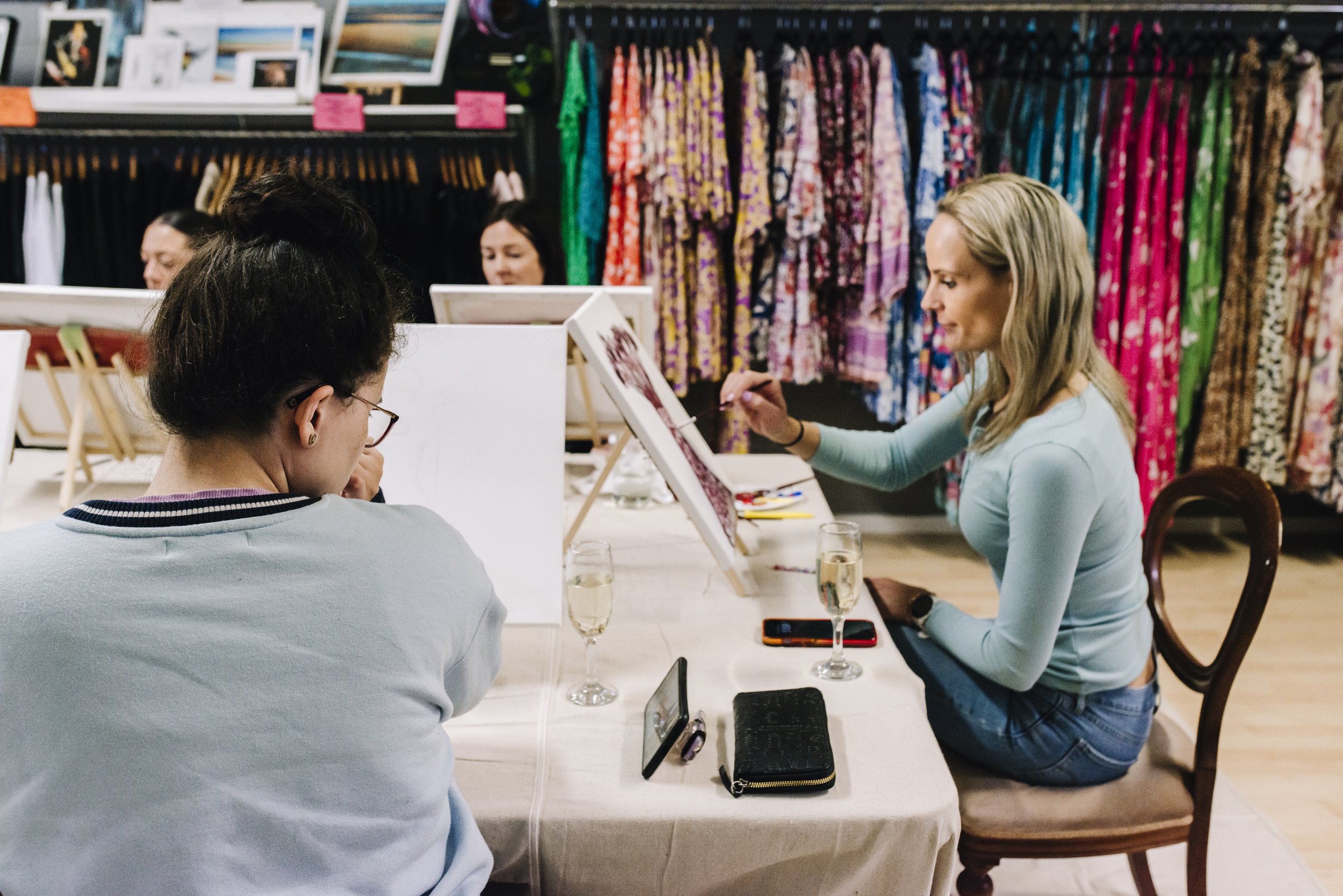 Women painting on canvases at a table inside a fabric store with colorful fabrics hanging in the background.