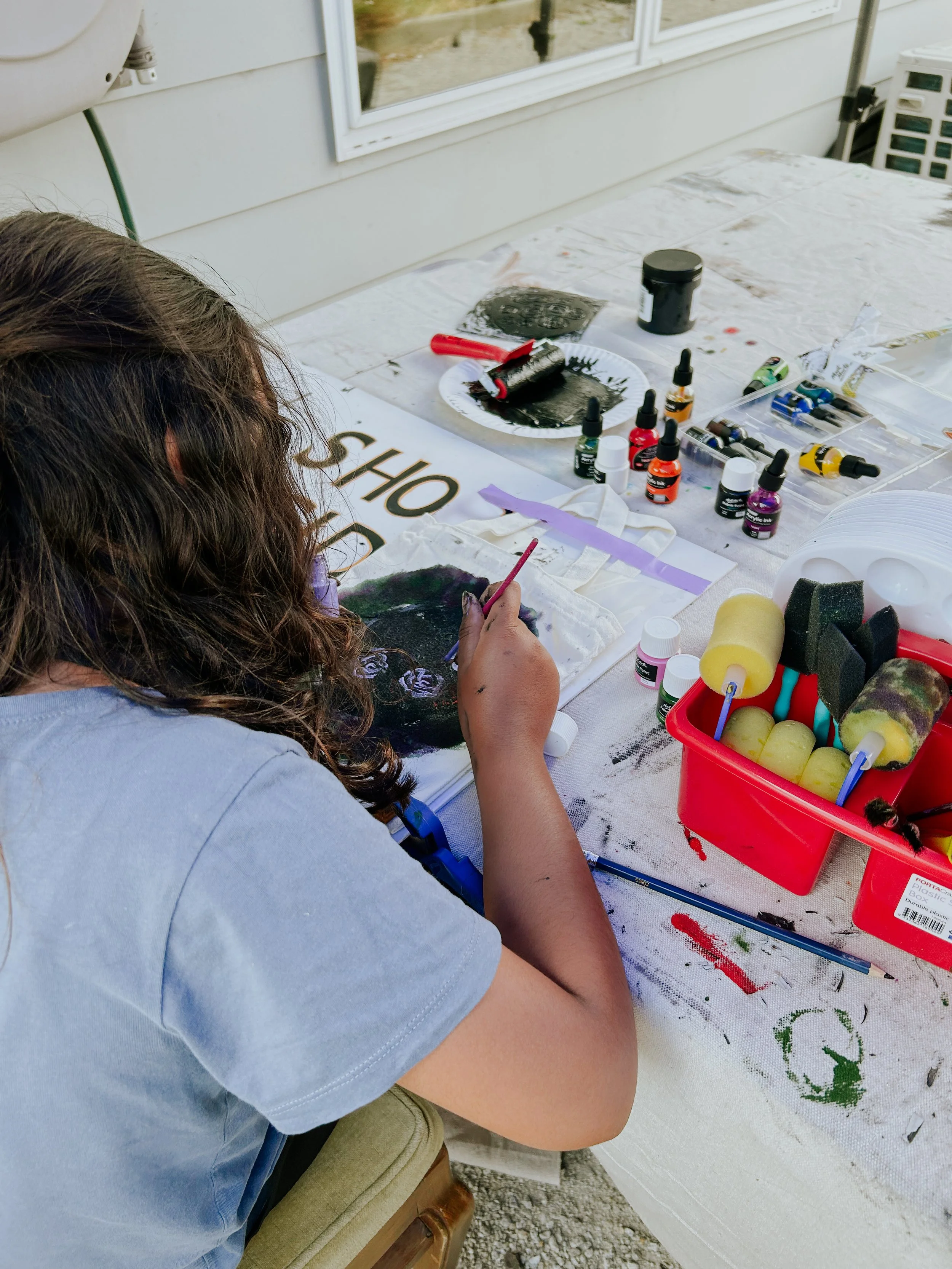 A person with long, curly hair is painting a black and purple design on a canvas using a small brush. The table in front is covered with various bottles of paint, sponges, and painting supplies. The setting appears to be outdoors or on a porch with a