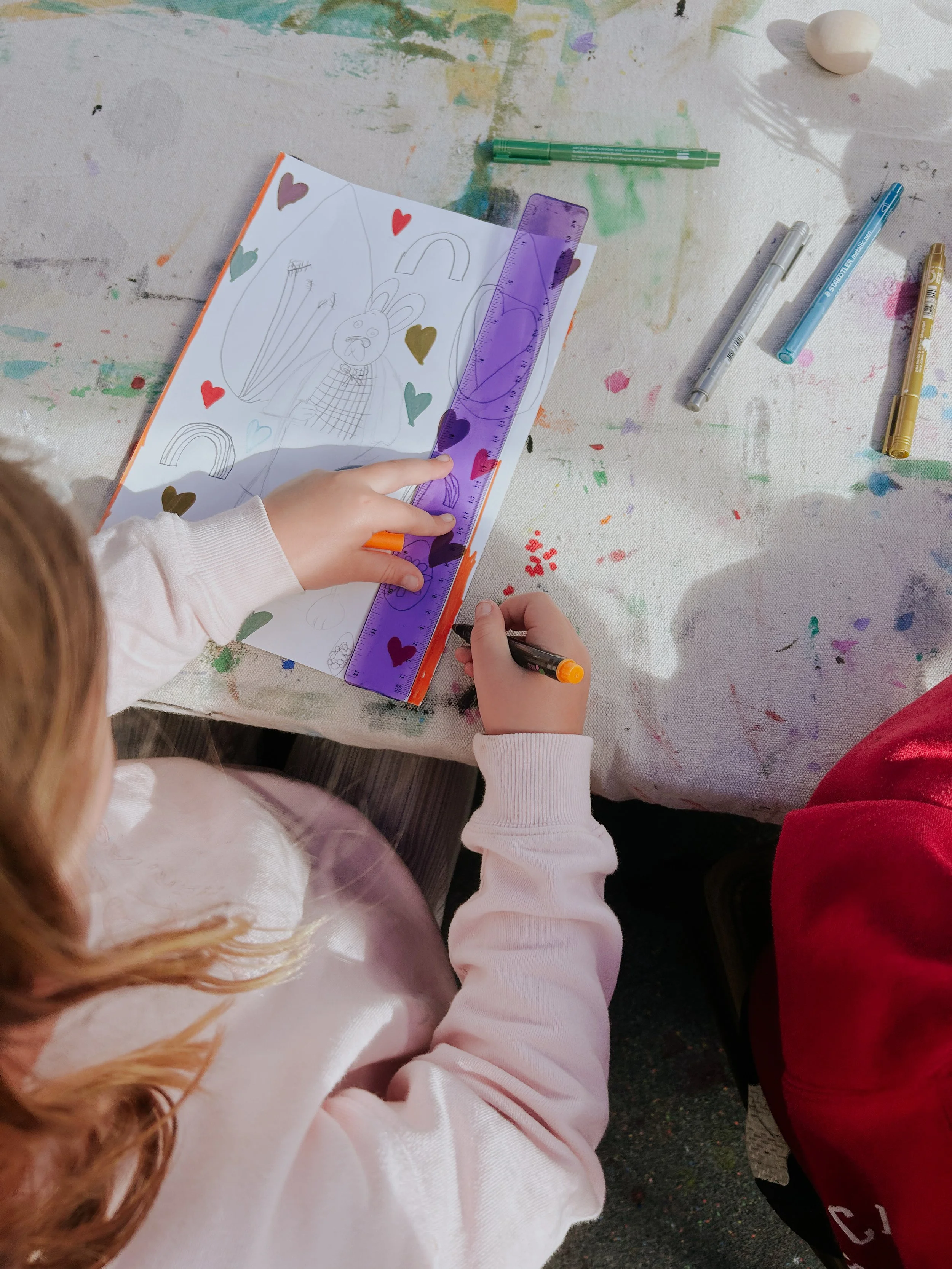 A child sitting at a table drawing with markers, ruler, and paper, which has a rabbit and rainbow illustration, on a paint-splattered table.
