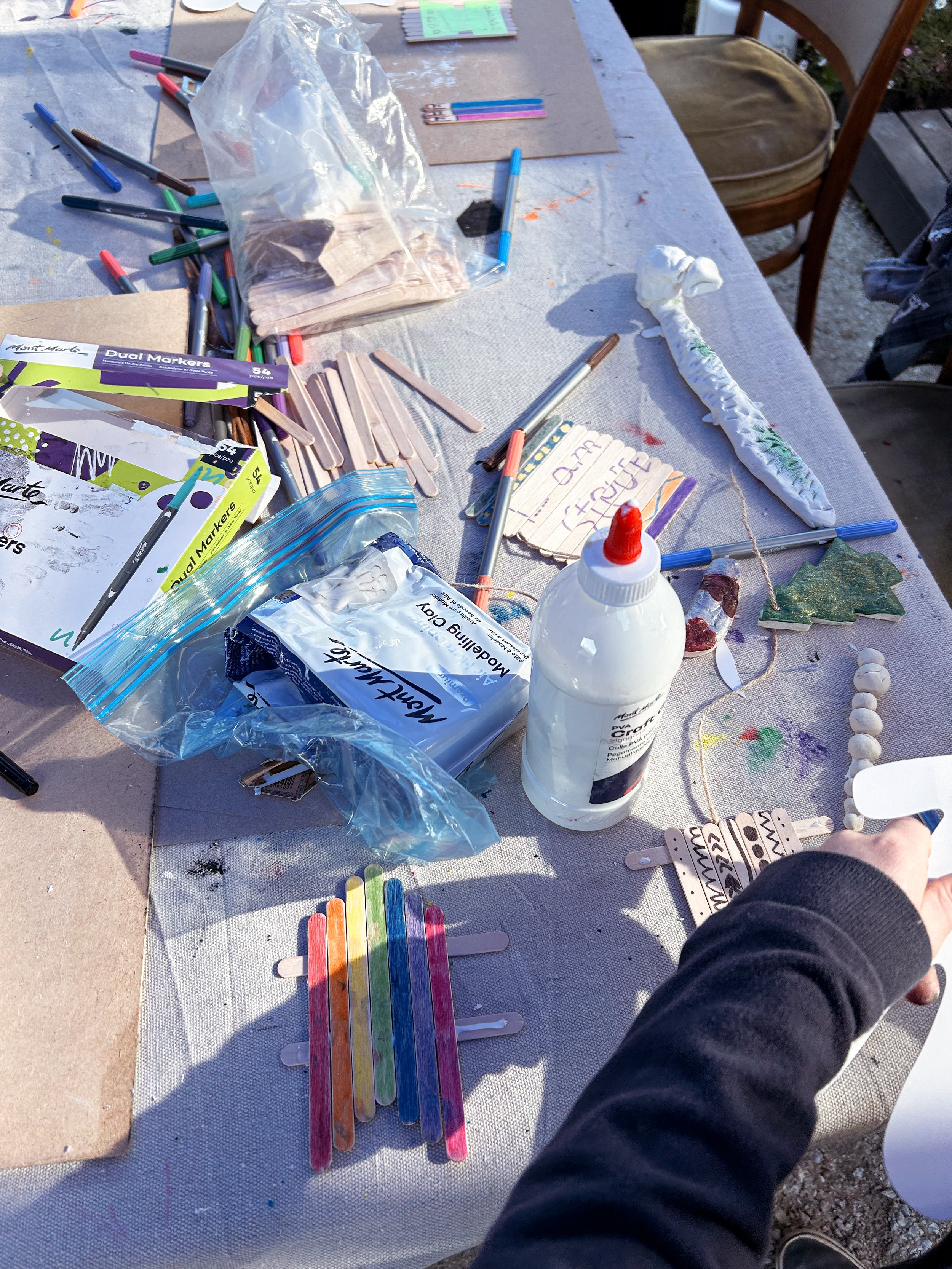 A table covered with arts and crafts supplies including markers, glue, popsicle sticks, popsicle stick snowflakes, foam ornaments, and decorated clothespins.