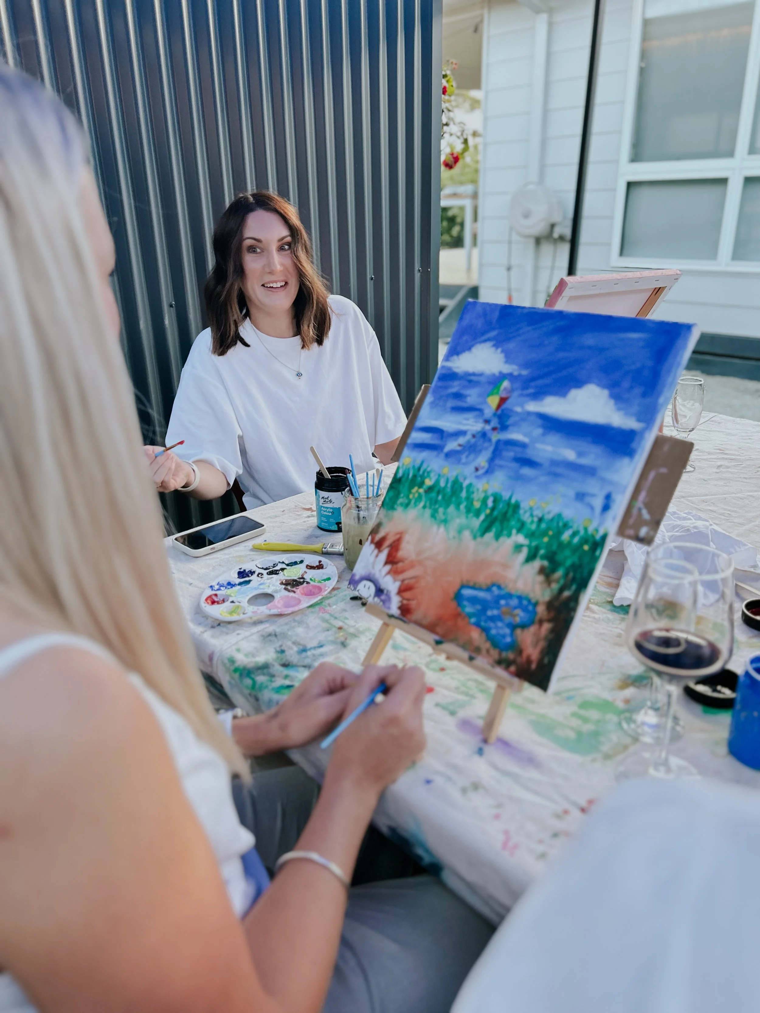 Two women painting a landscape on canvas outdoors, with one woman smiling and engaging in conversation.