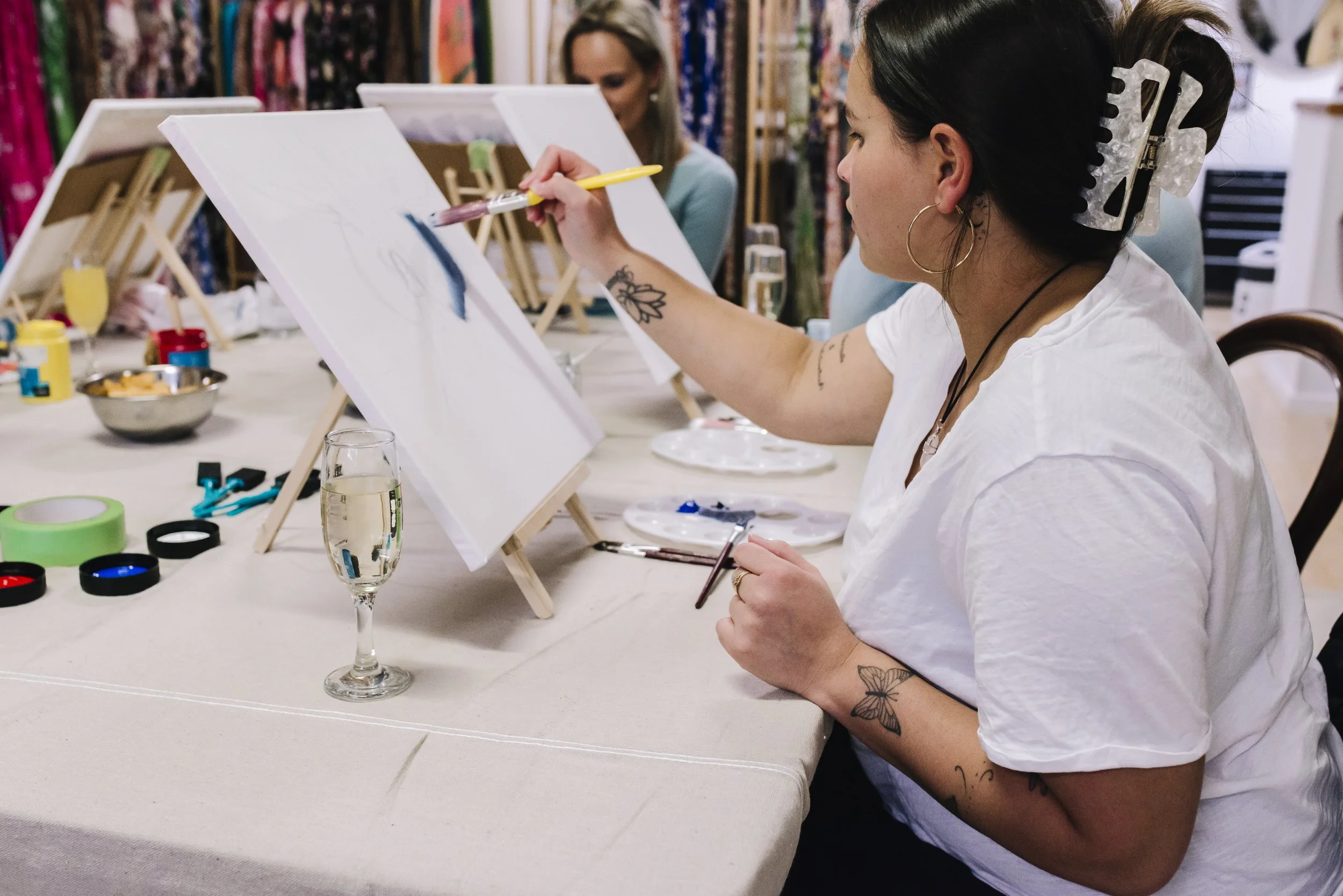 Women painting on canvases at an art class, sitting at a table with painting supplies and drinks, with colorful fabric patterns in the background.
