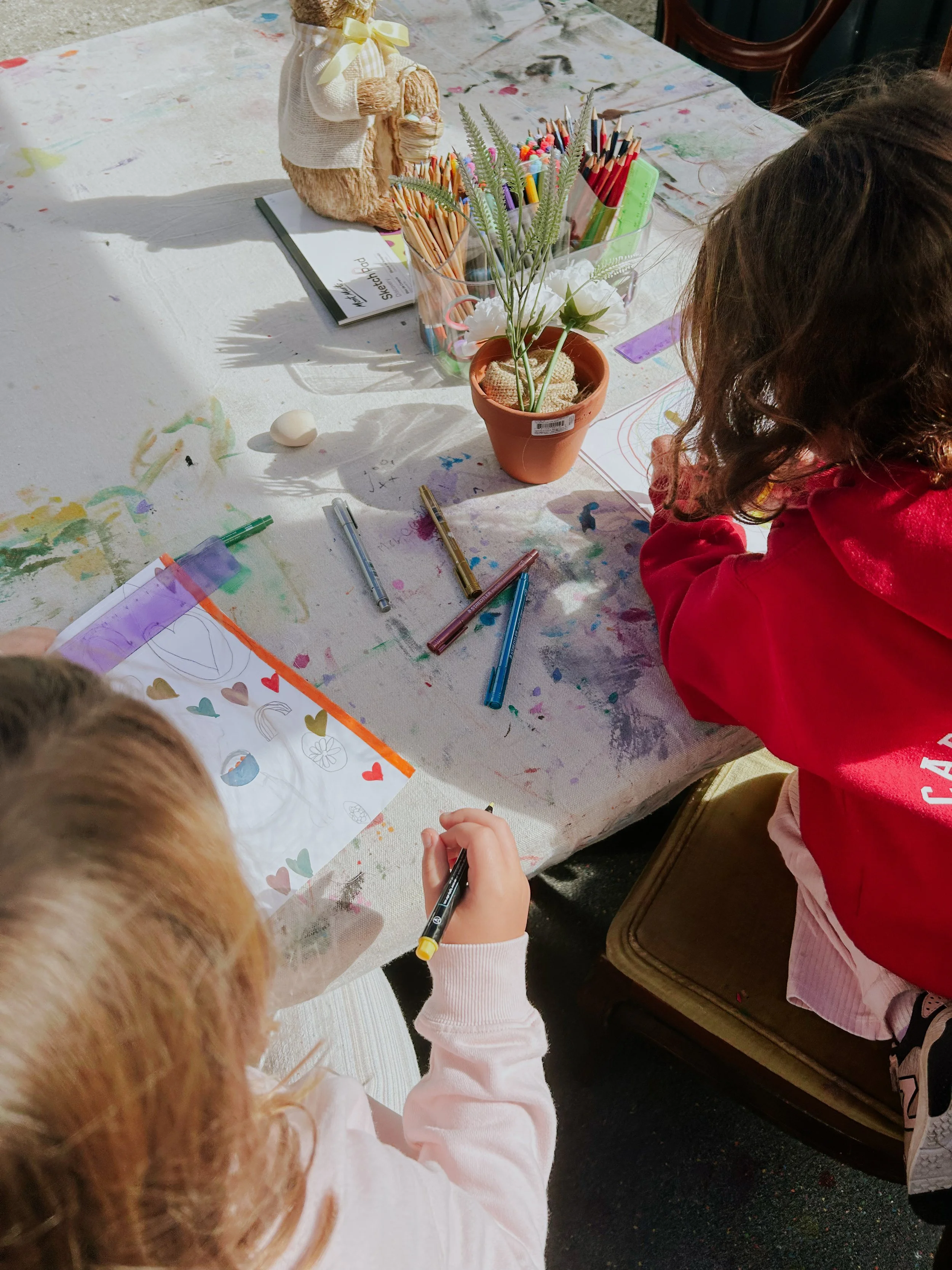Two children sitting at a table engaged in arts and crafts, with drawing supplies, paper, and decorative items on the table.