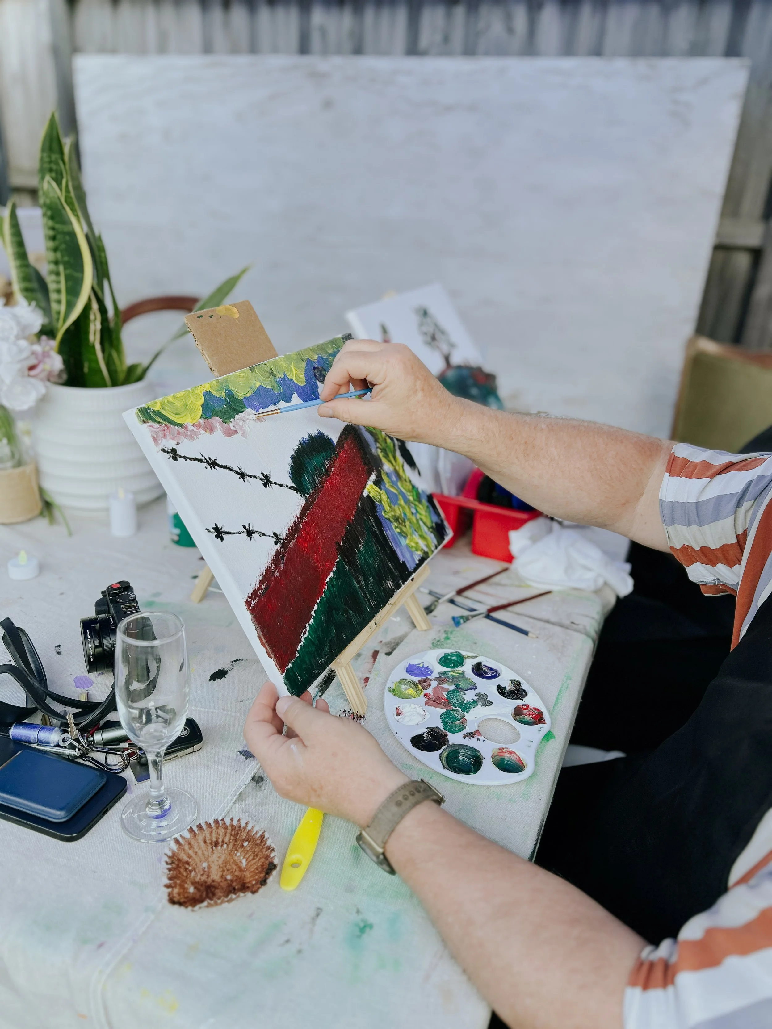 Person painting an abstract landscape on a canvas set on a small easel, with art supplies, a palette, and plants on the table.
