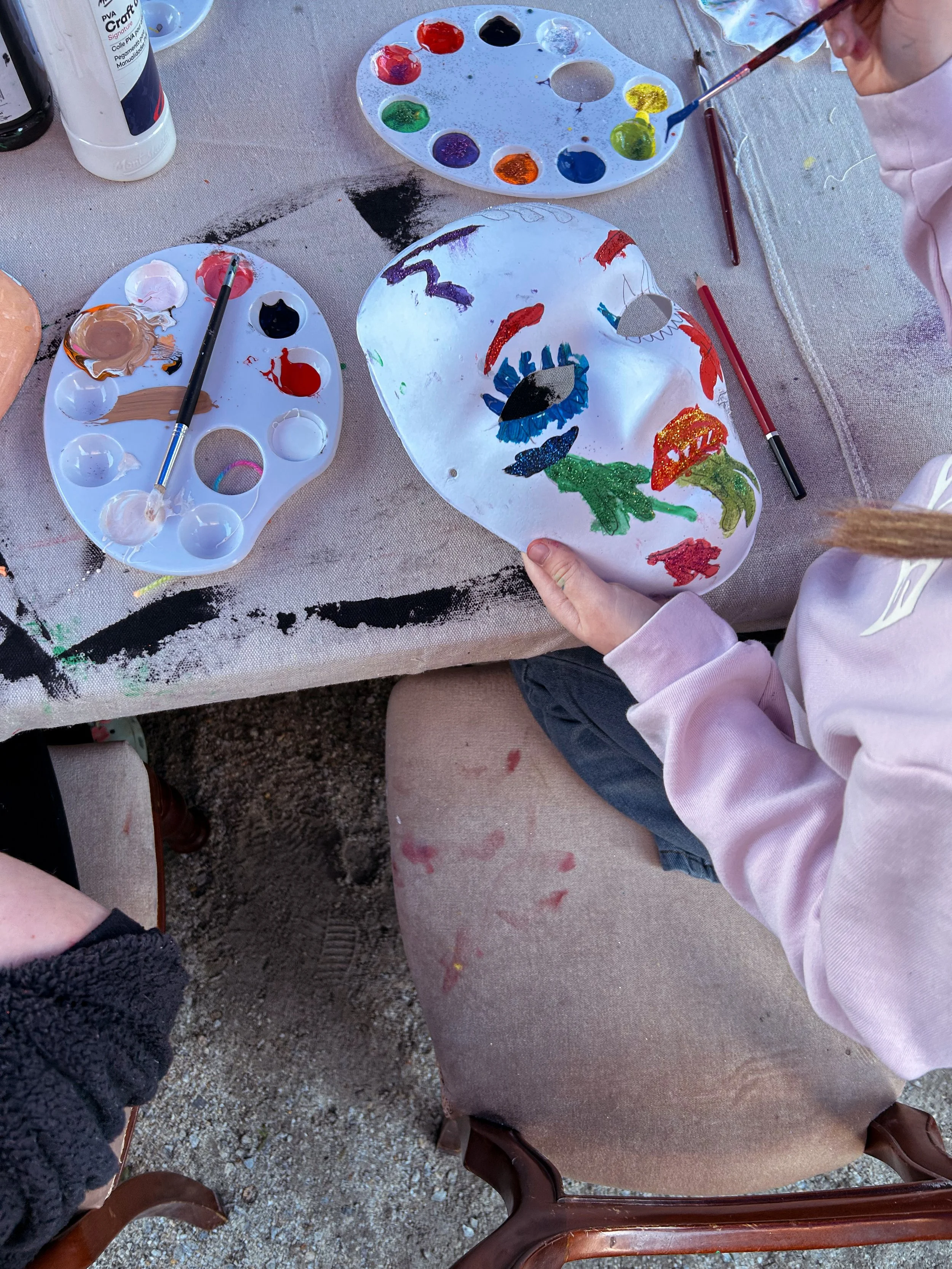 A child holding a painted white mask with colorful designs, sitting at a table with paint palettes and brushes, with some paint on their hands and clothes.