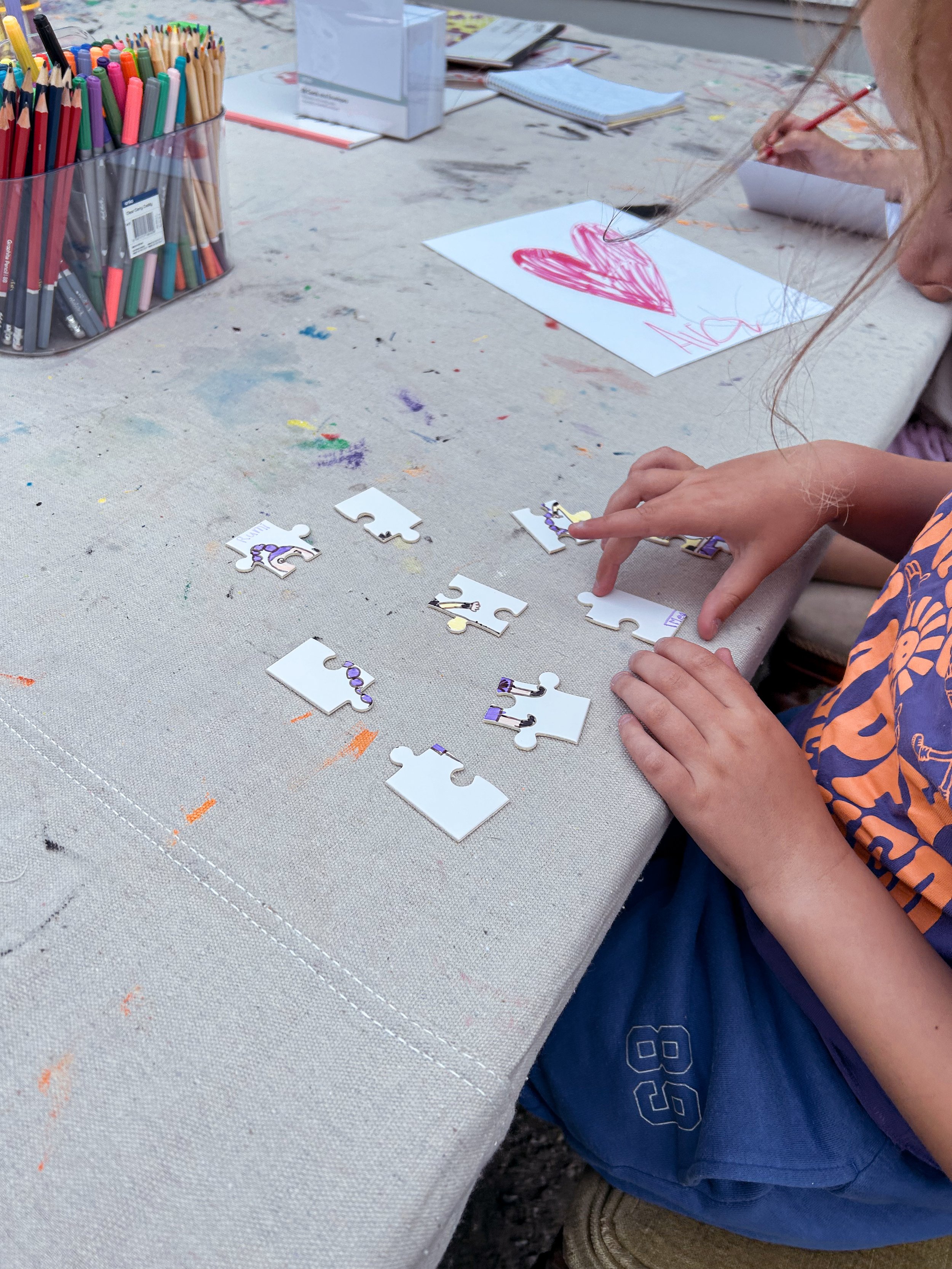 Child assembling a puzzle with unicorn designs on a table covered in paint stains, with a drawing of a pink heart and other art supplies in the background.