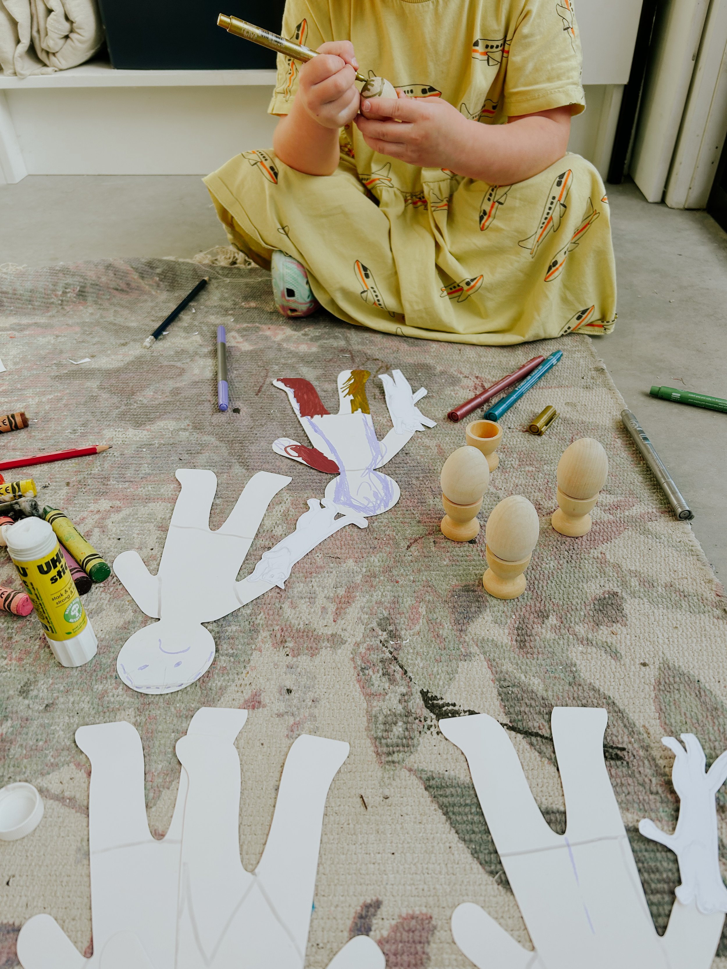 Child in yellow pajamas sitting on the floor and working on a paper craft project with paper cutouts, markers, glue, and wooden egg-shaped objects scattered on a rug.