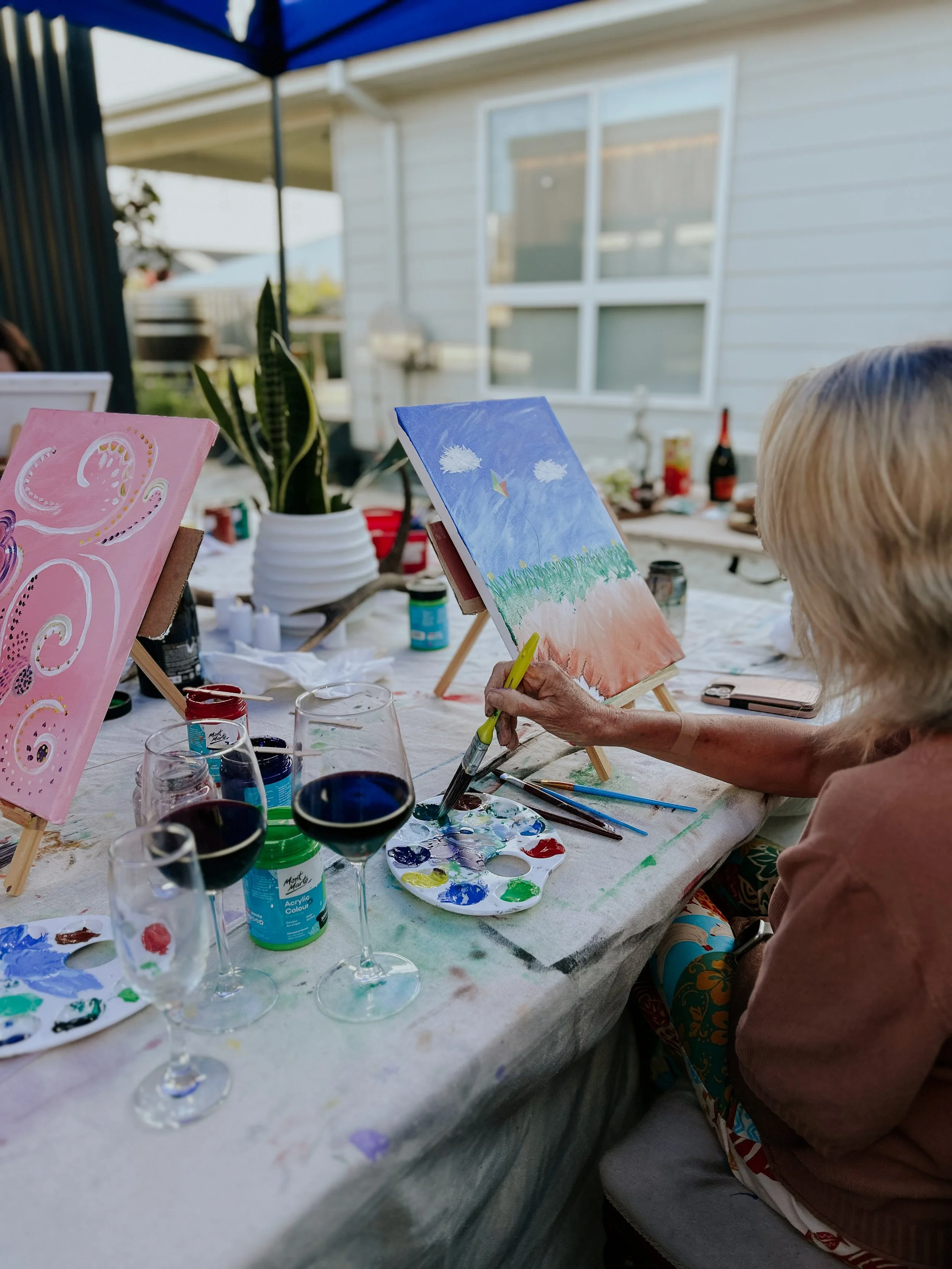 Woman painting a landscape on canvas during outdoor social gathering, with various paints, brushes, wine glasses, and canvases on the table.