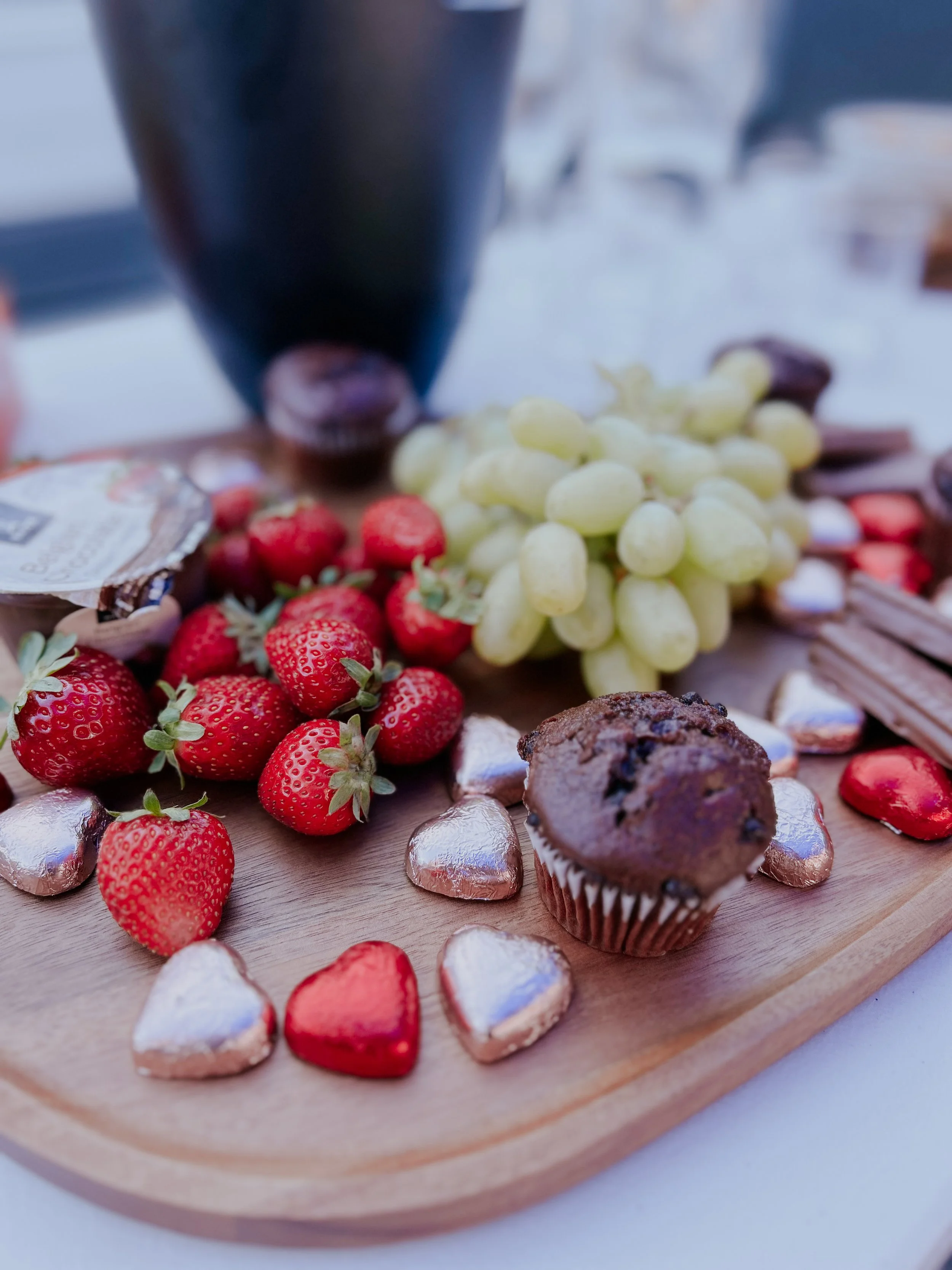 A wooden serving board with strawberries, white grapes, a chocolate muffin, and small heart-shaped chocolates.