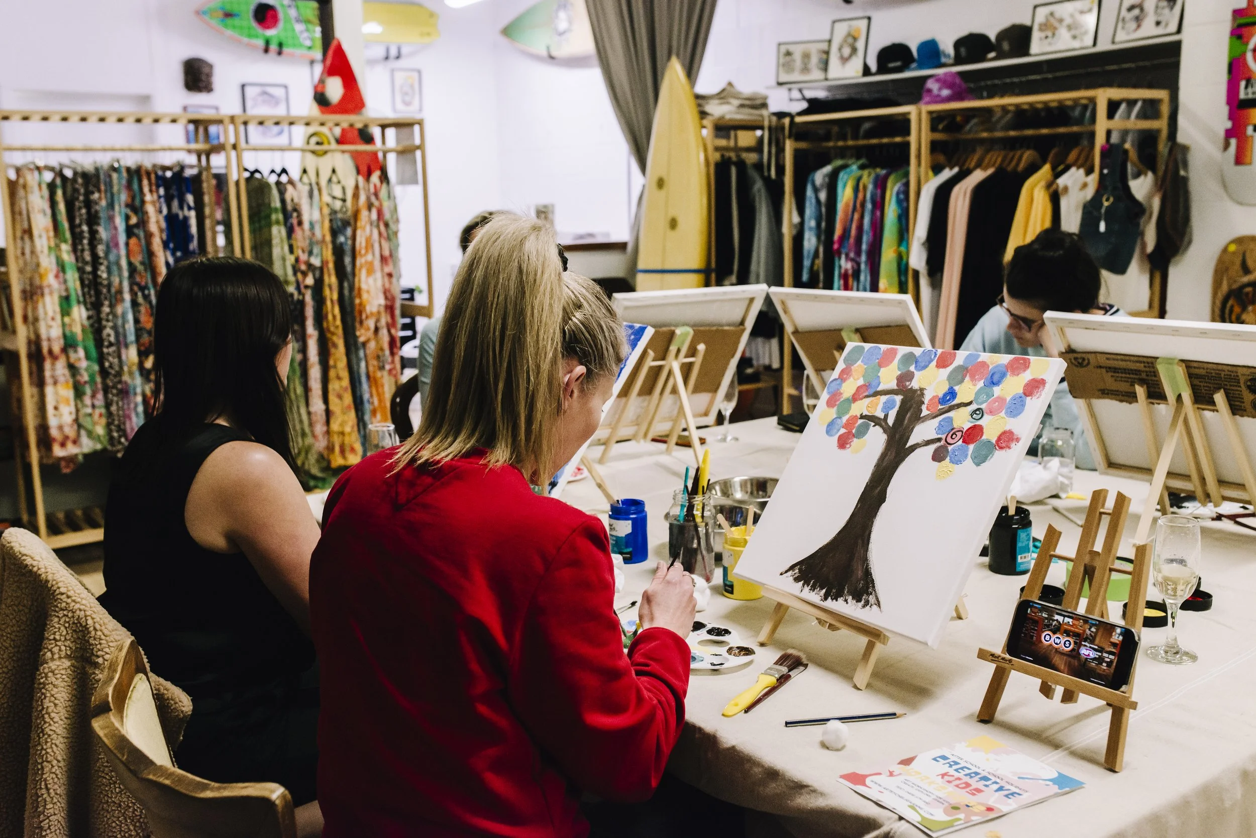 People participating in a painting class in a room filled with colorful clothing and art displays, with a canvas of a tree with multicolored leaves.
