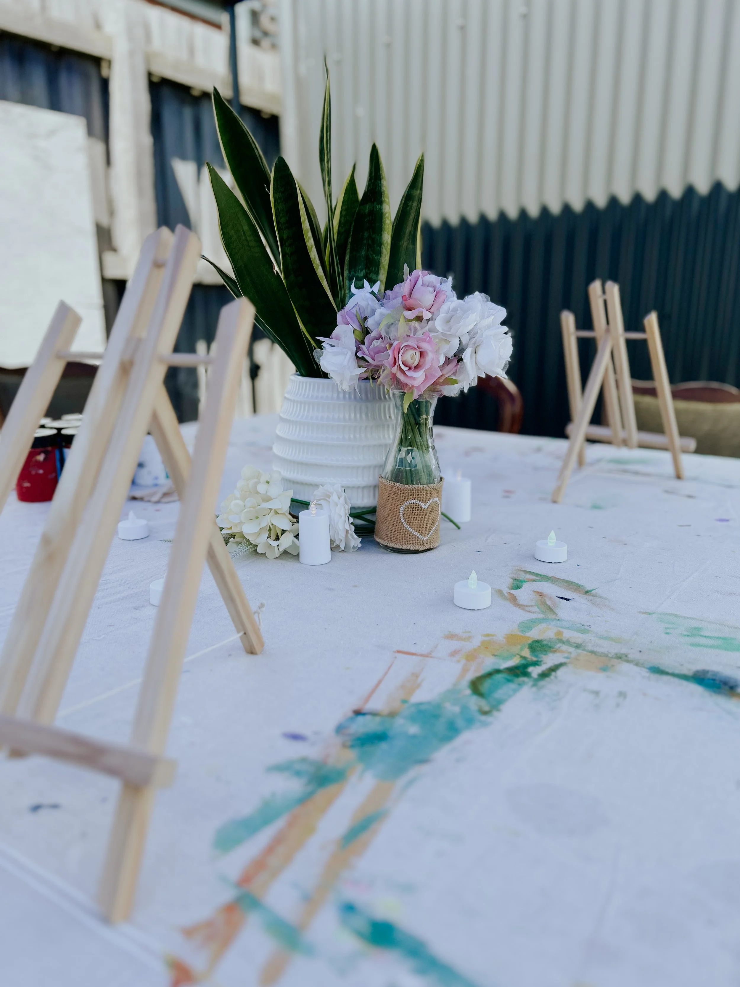 Table with a floral centerpiece, small candles, and wooden easels, outdoors with a canvas and a fence in the background.