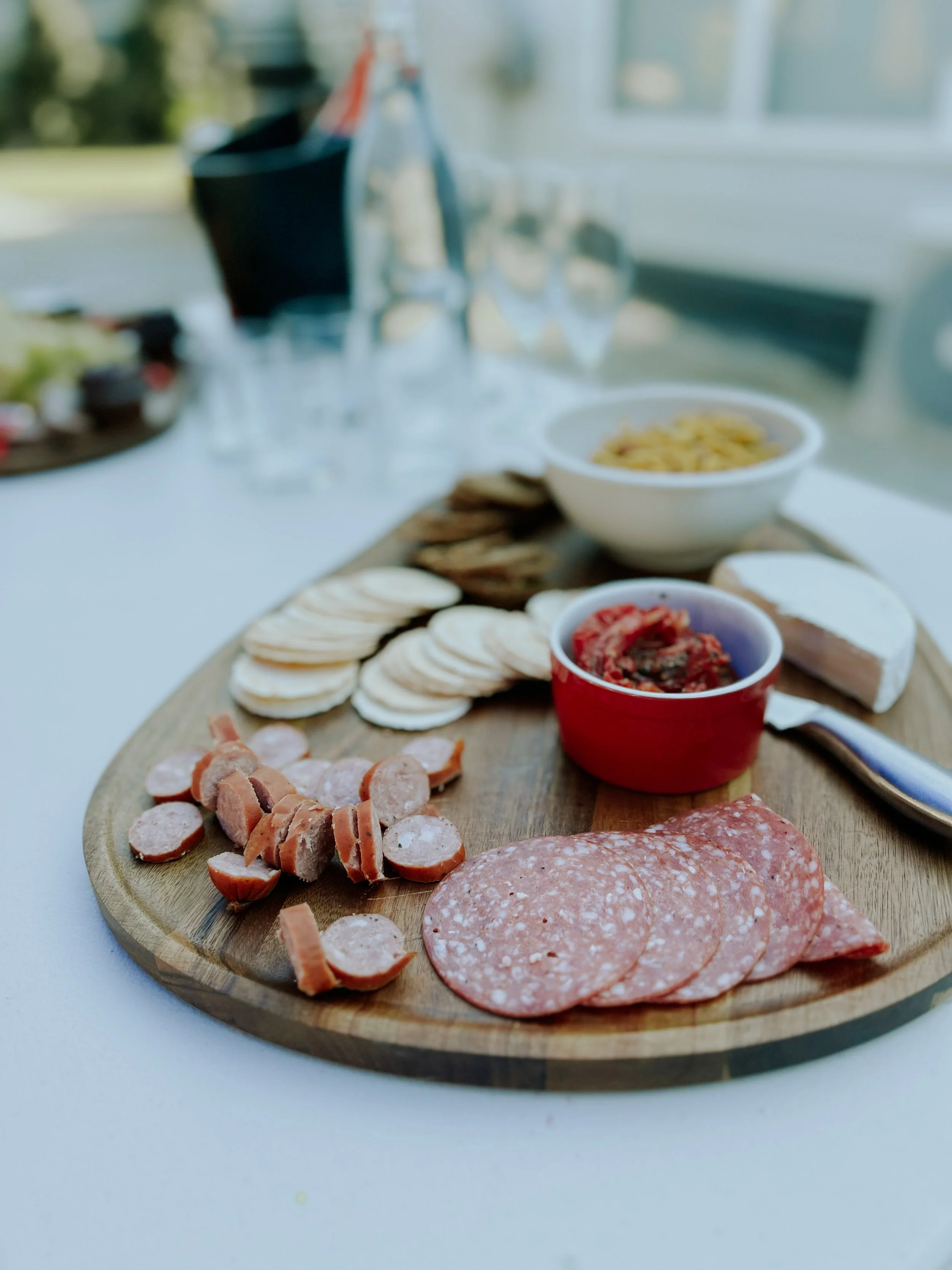 Charcuterie board with sliced meats, crackers, and jams on a wooden platter, with bowls of nuts and olives in the background.