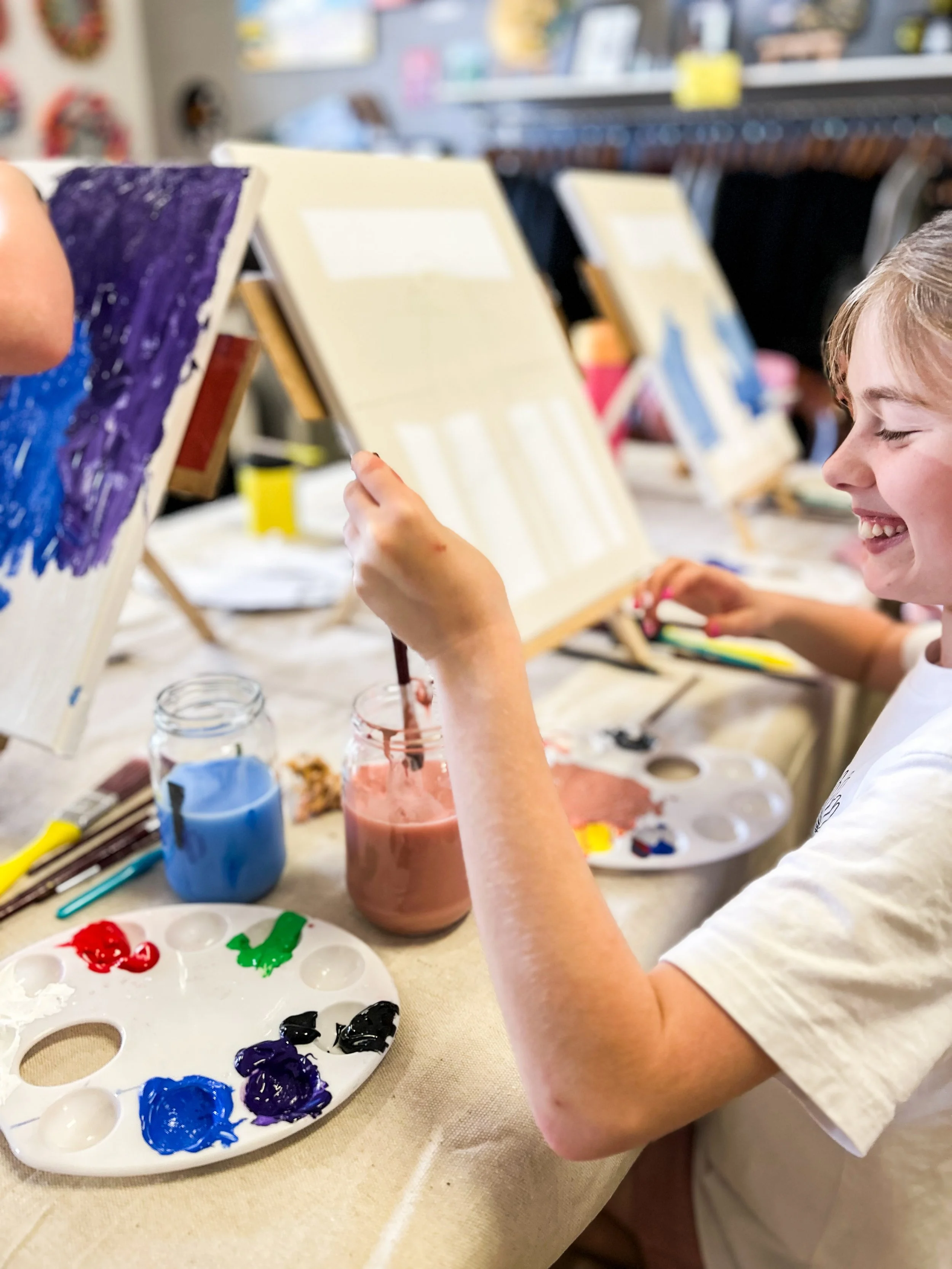 A young girl is painting on a canvas at an art class, with a big smile on her face. Various paint colors and painting supplies are on the table in front of her.