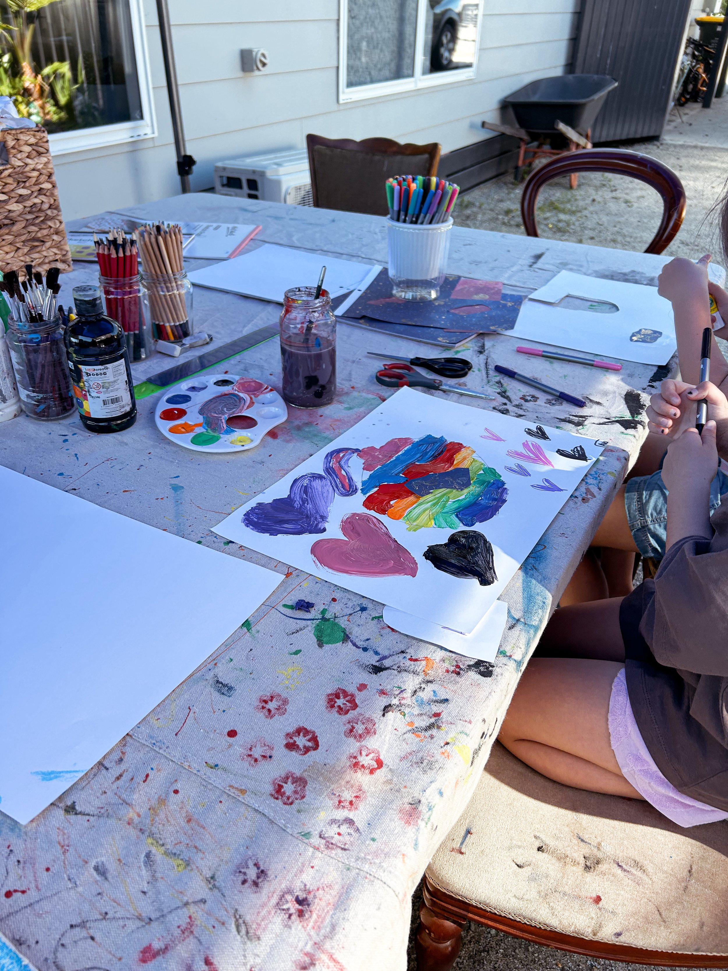 Children painting colorful hearts on paper at an outdoor table with art supplies, in a backyard setting.