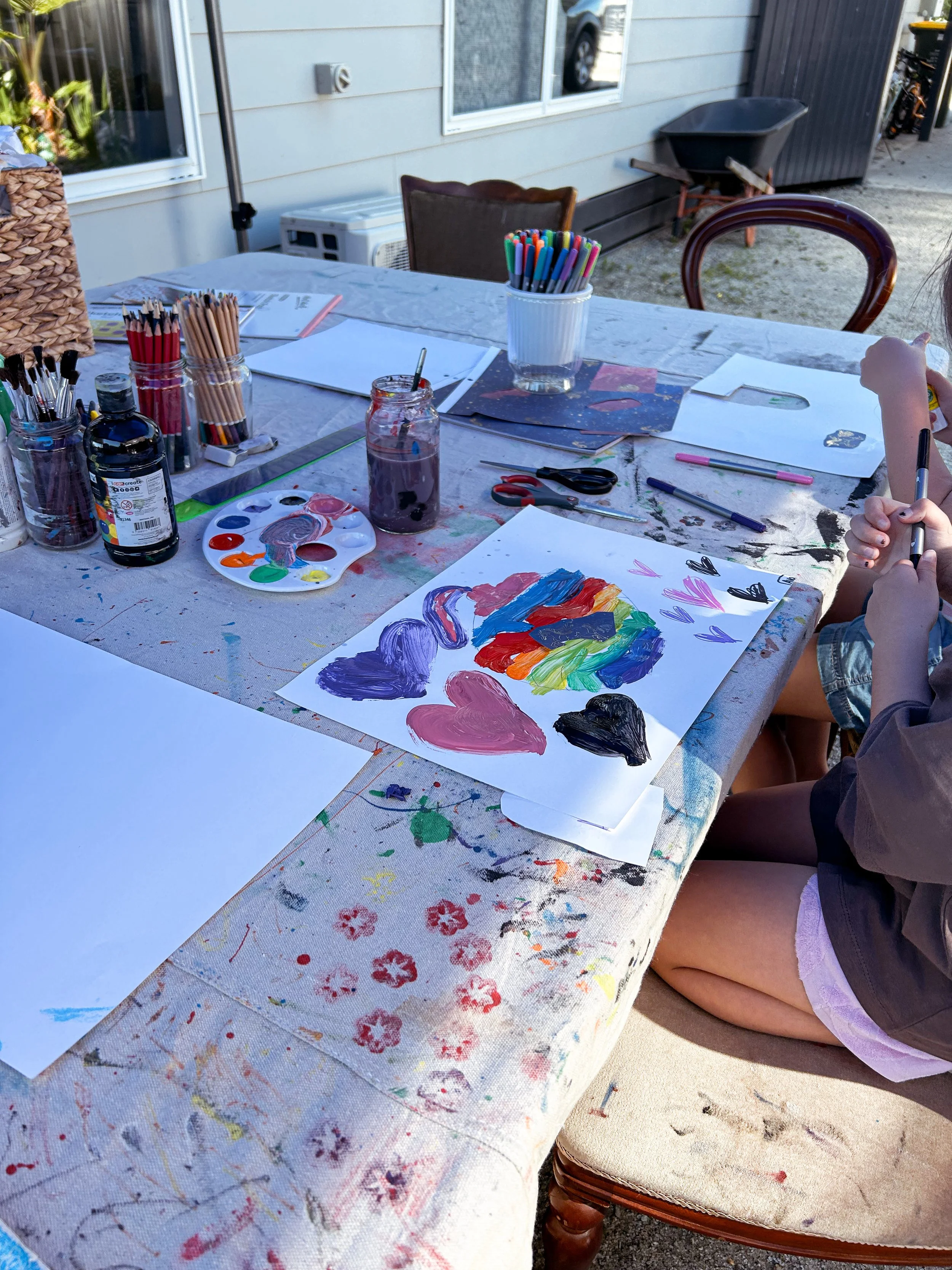Children creating colorful heart-shaped paintings at an outdoor art table with paint supplies.