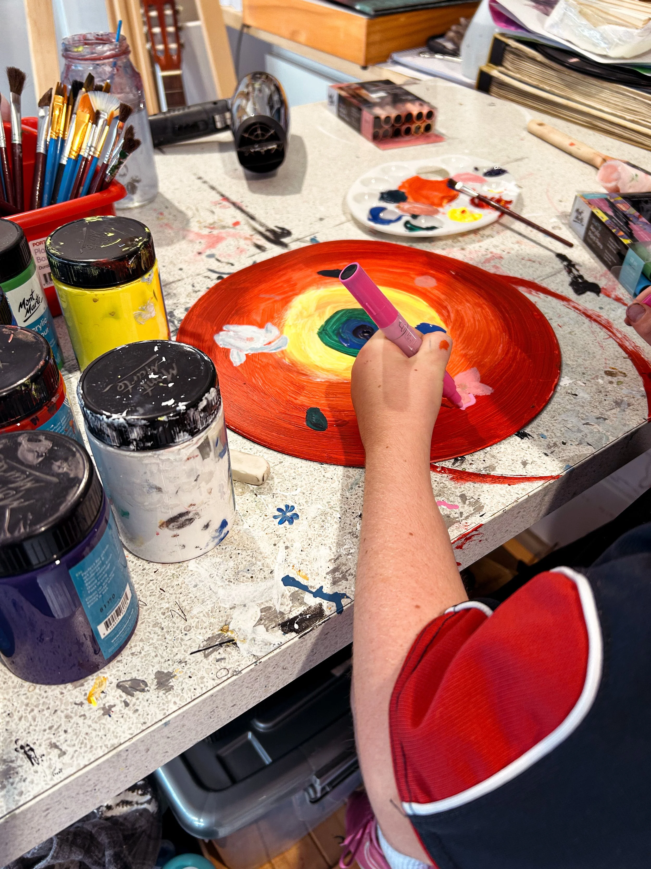 A person painting a circular abstract artwork with orange, yellow, green, blue, and white colors using a pink marker, on a cluttered art table with paint jars, brushes, and art supplies.