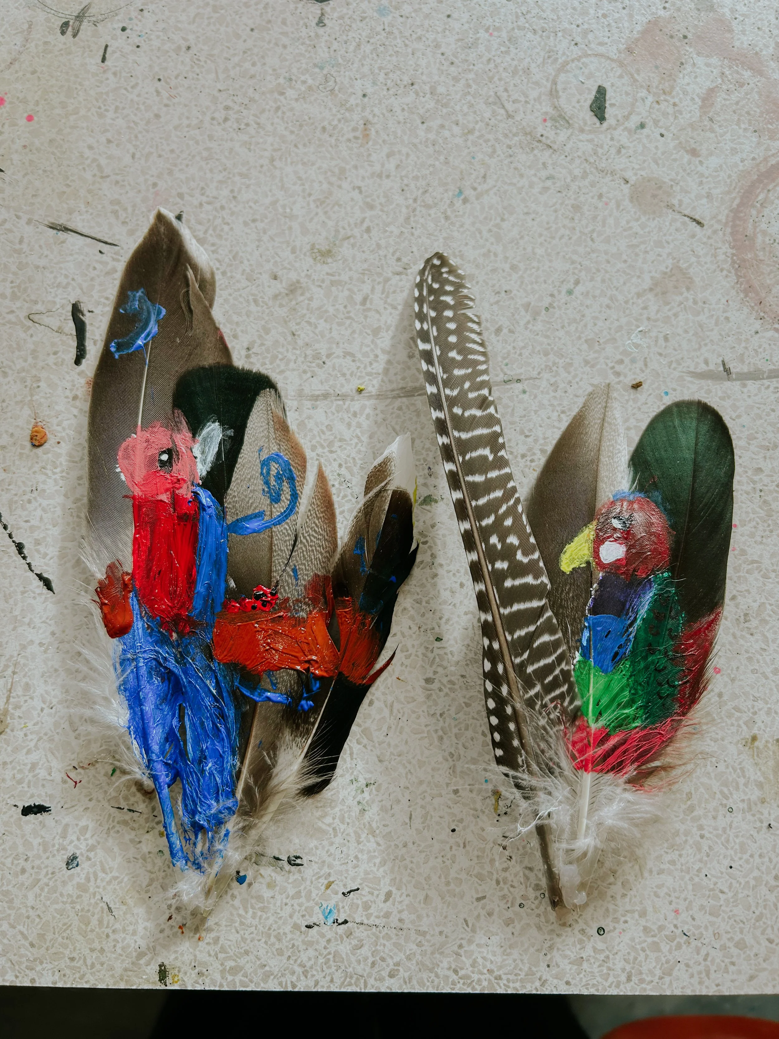 Four feathers with paint splatters on them, likely used for art or craft projects, resting on a marbled surface with scattered small paint spots.