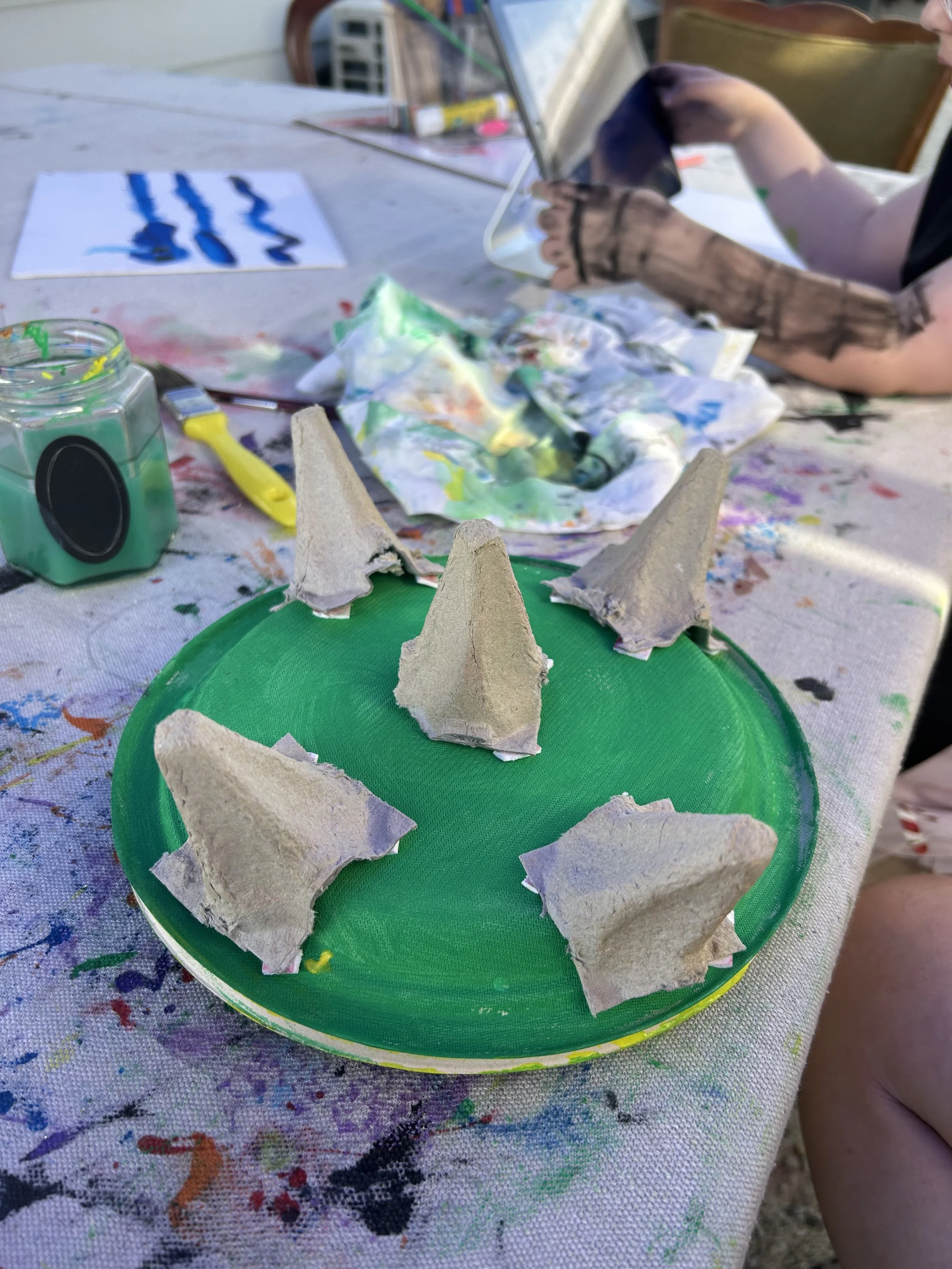 Painted green plate with five clay spikes arranged to resemble a face with a toothy smile, surrounded by art supplies on a table covered with colorful paint stains.