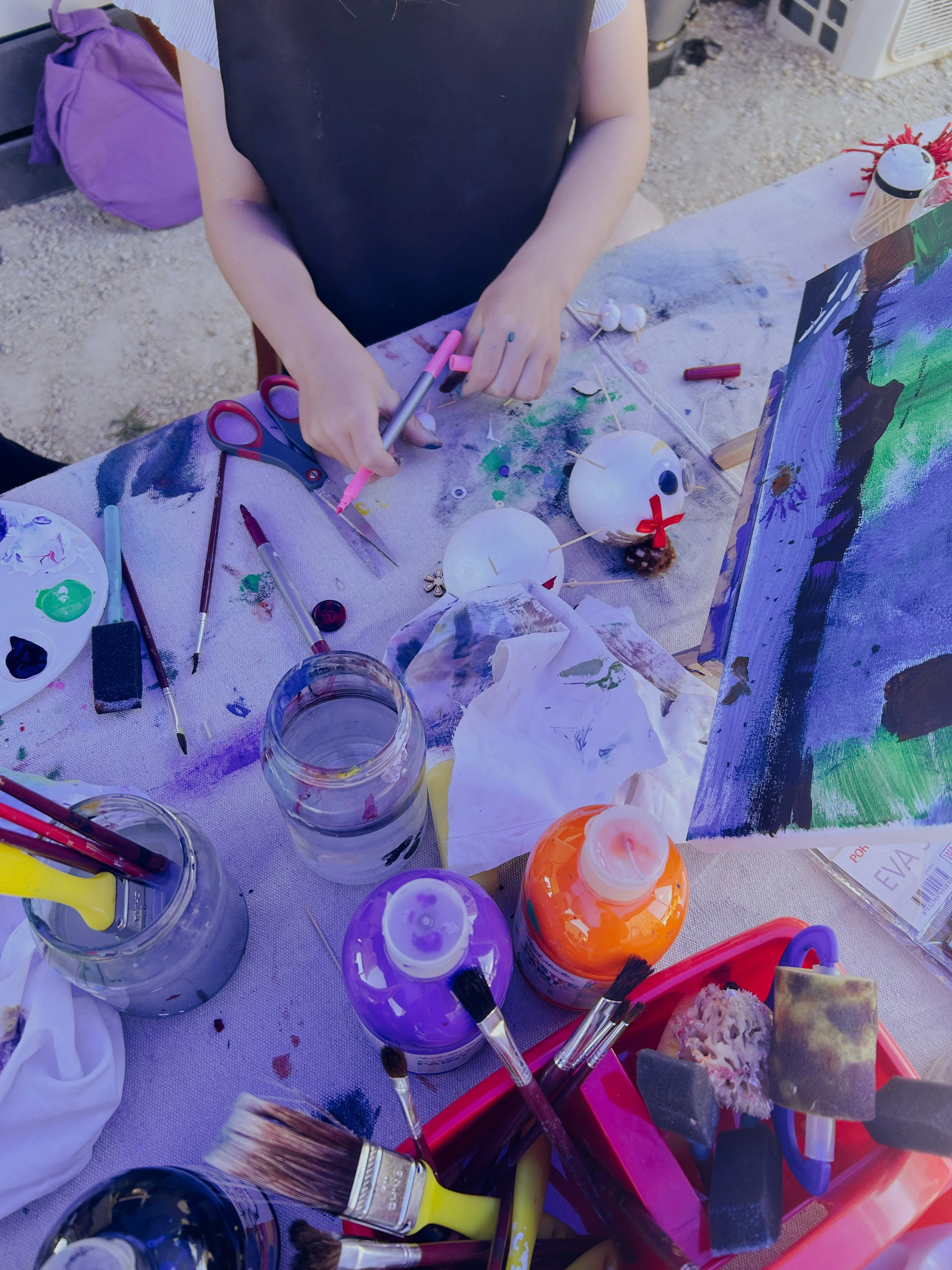 A person painting decorative ornaments at an outdoor art station, surrounded by paint bottles, brushes, scissors, and art supplies.