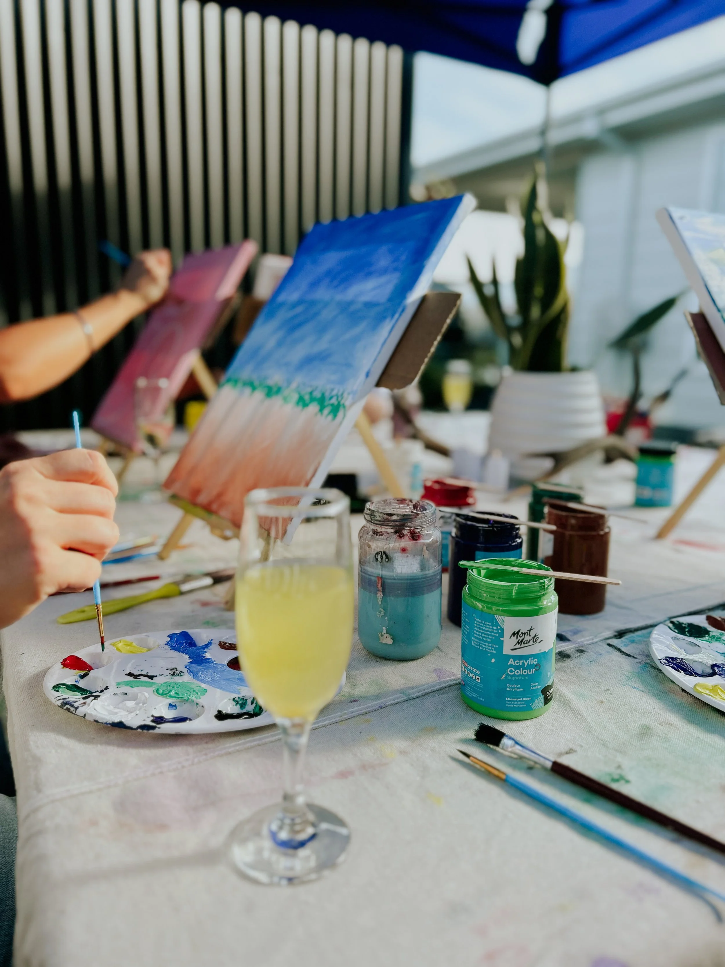 People painting on canvases set up on small easels in a bright room with large windows, various jars of paint, brushes, and a glass of champagne or white wine on the table.
