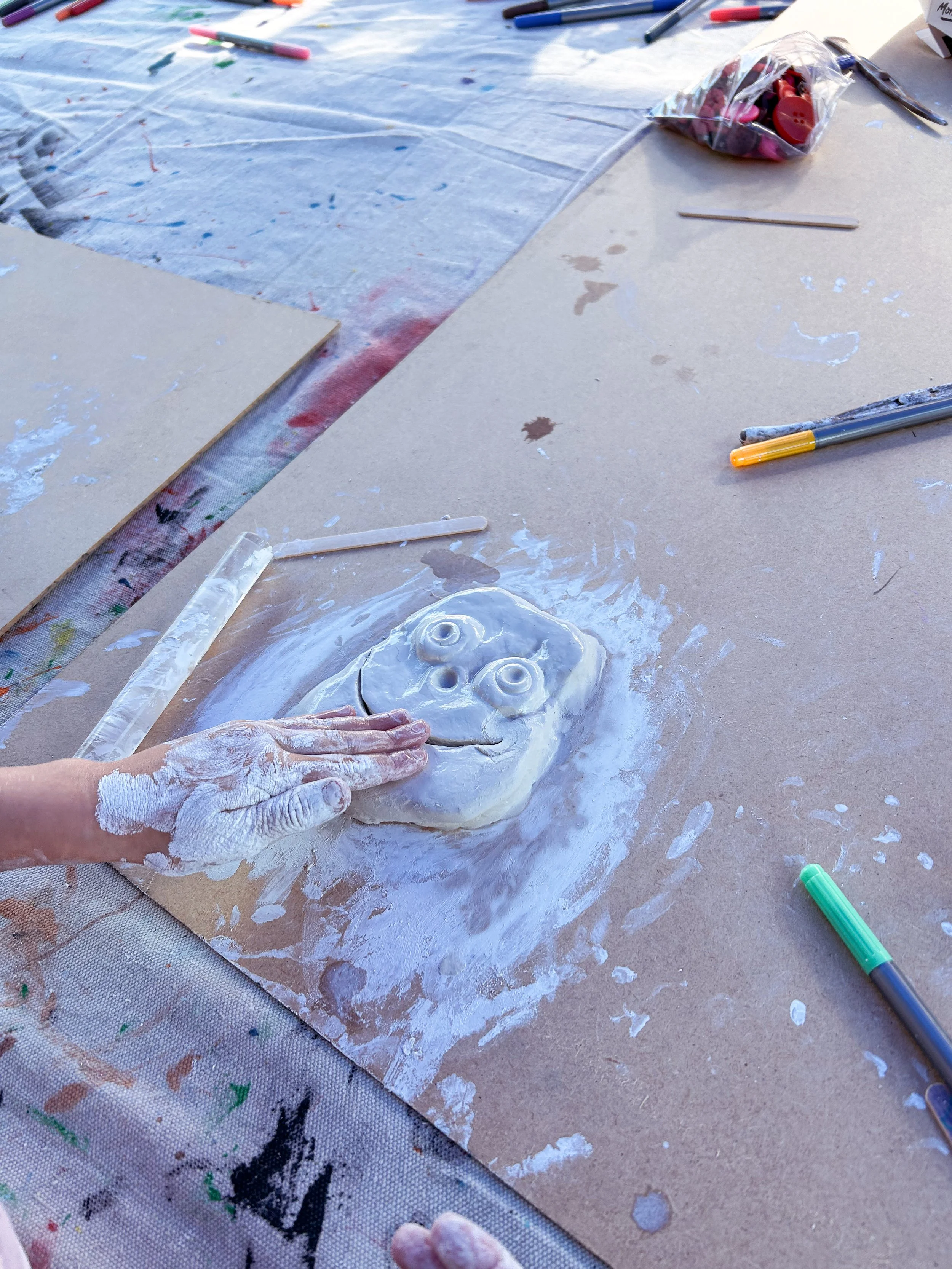 Child creating a smiling face with large eyes using white clay on a brown paper surface, surrounded by art supplies like markers, paint, and brushes.