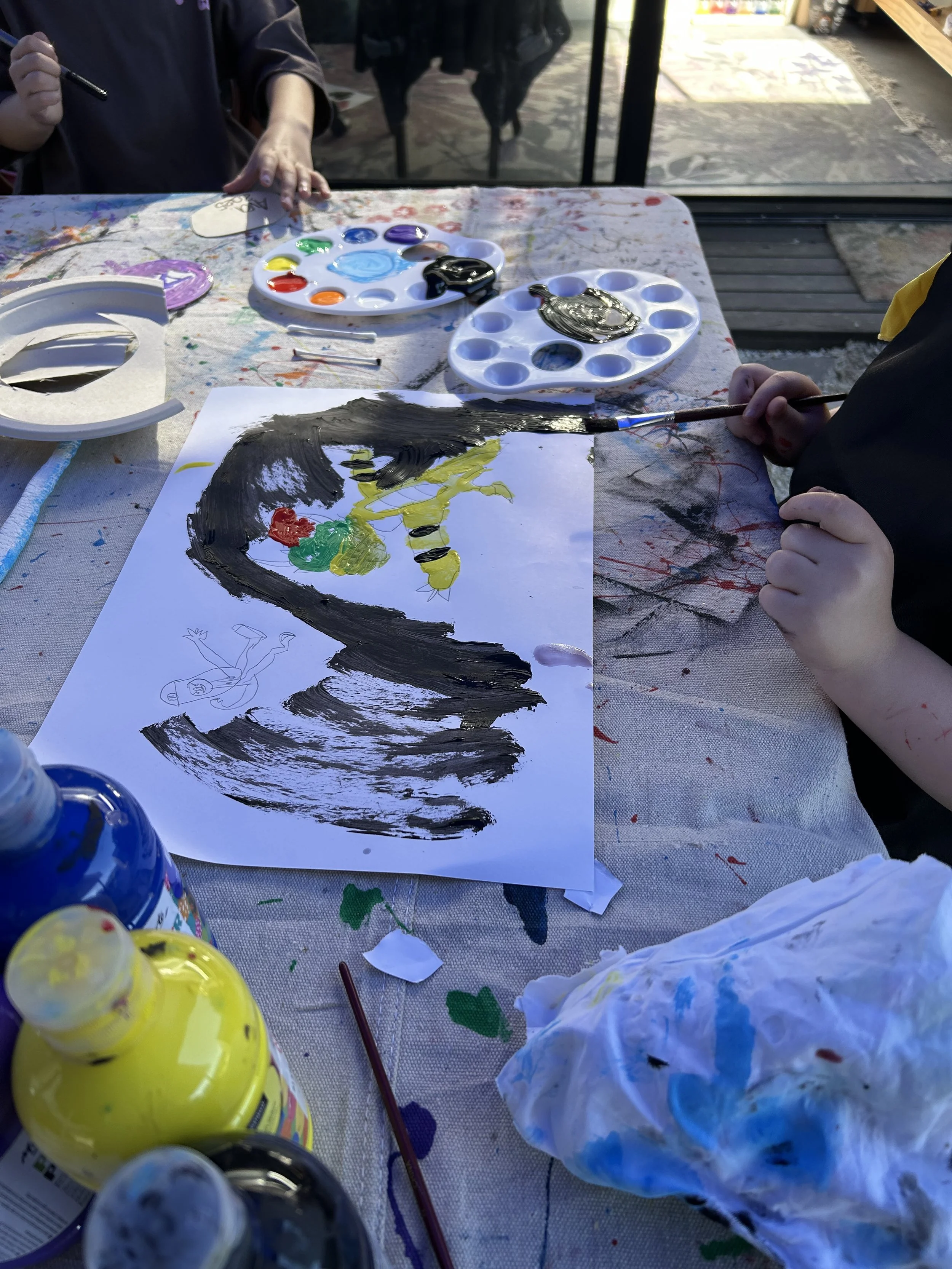 Children painting a picture using black, yellow, green, and red paint on a white sheet of paper, with paint palettes and bottles of paint on a paint-splattered table.