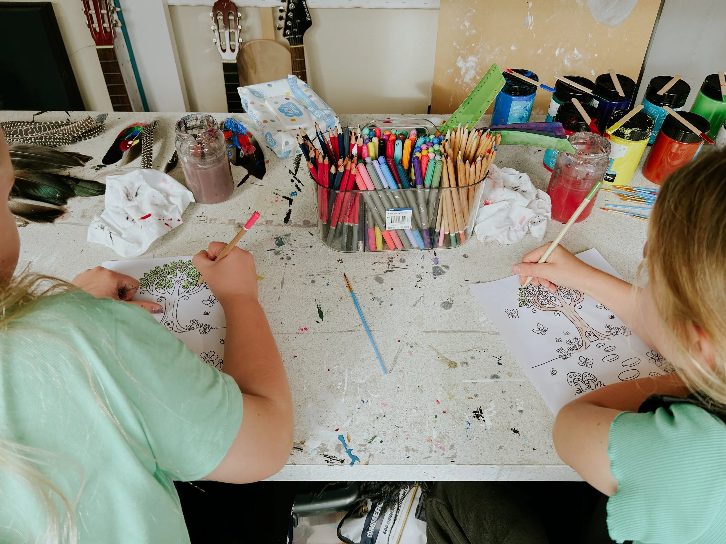 Children coloring pictures of trees at a table with art supplies, including colored markers, paint jars, and paper towels.
