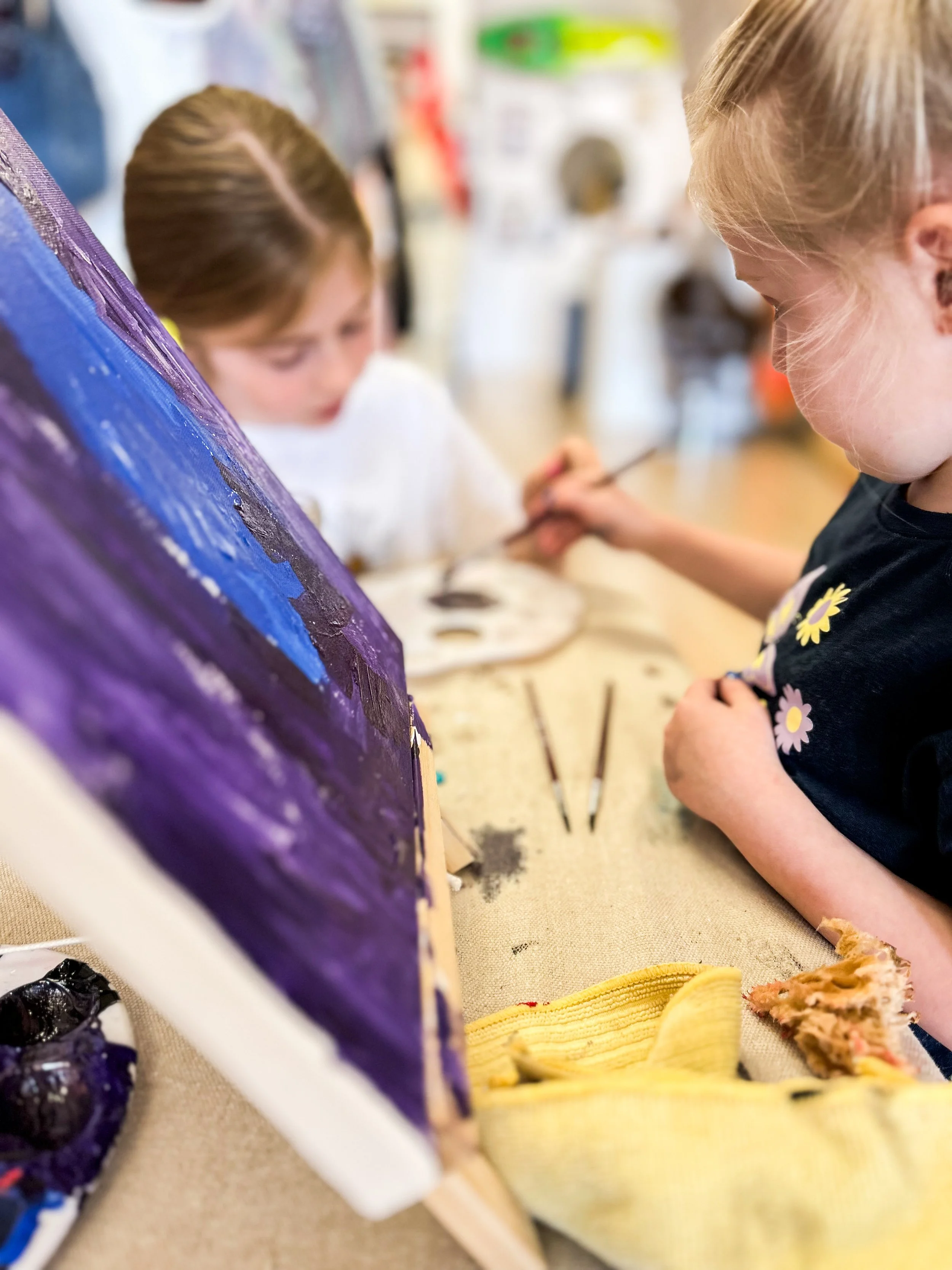 Two young children painting on a canvas at a table in an art classroom. One girl is painting while the other looks on.