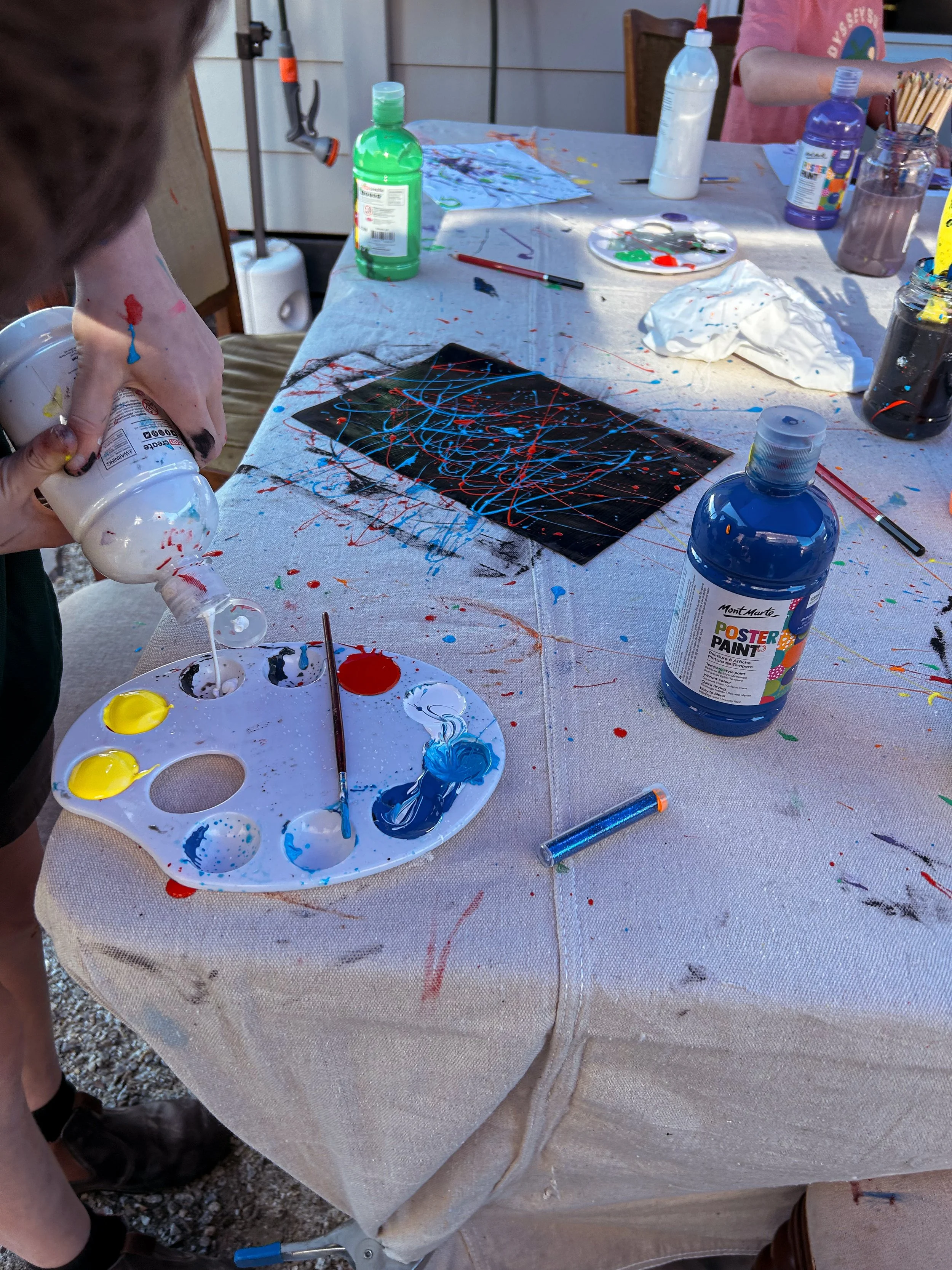 A table covered with splattered paint, paint bottles, a palette with yellow, red, and blue paint, paintbrushes, and a canvas with an abstract splatter painting. A person is pouring white paint from a bottle onto the palette.