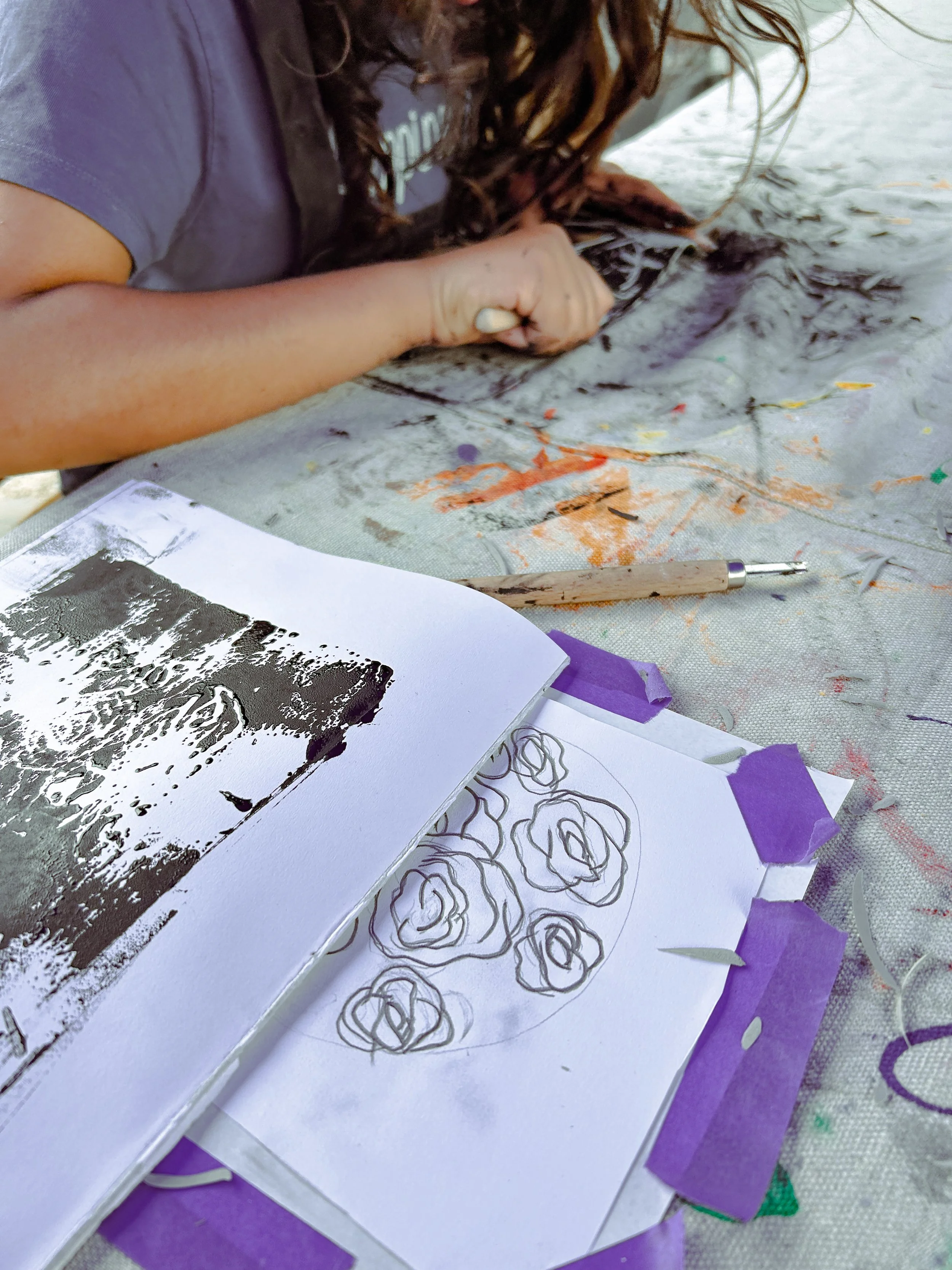 A person with curly hair is working on an art project outdoors, using a pen on a canvas. There are sketches of flowers and a reference photo on paper, with art supplies and purple tape on a cloth surface.