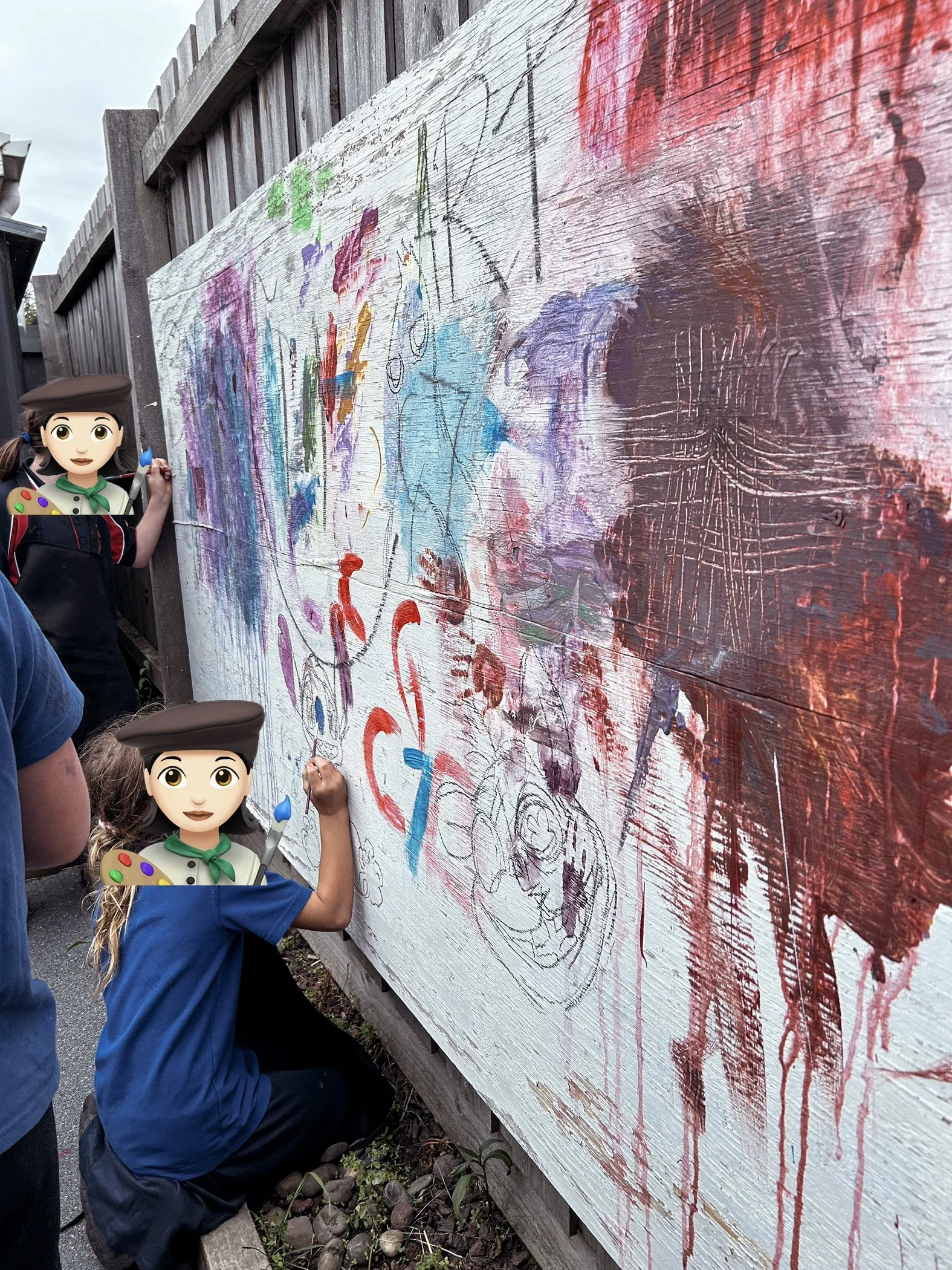 Children creating artwork on a large outdoor wooden wall, with colorful paint and sketches.