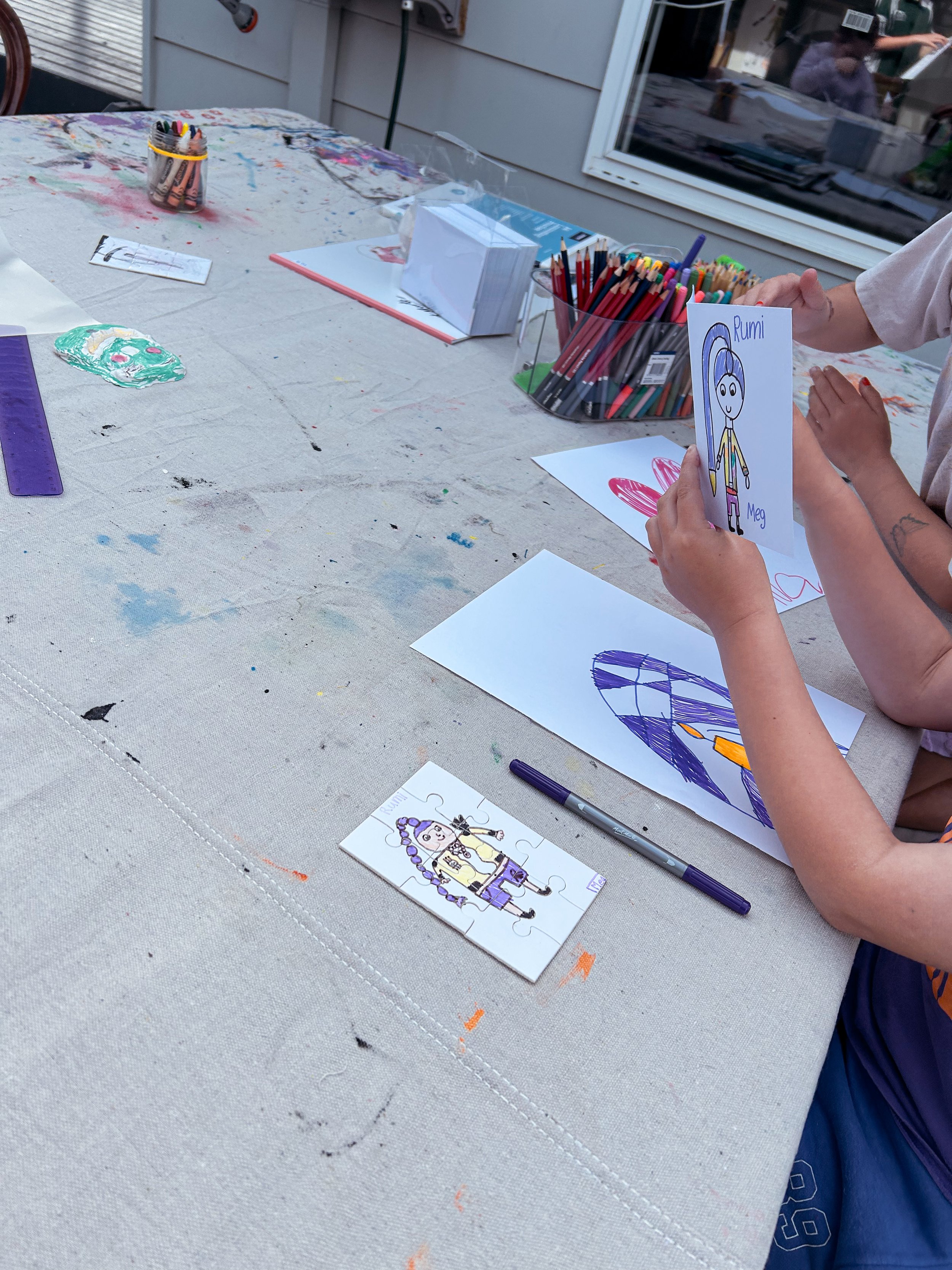Children at a table with coloring sheets, markers, and arts and crafts supplies outside on a patio.