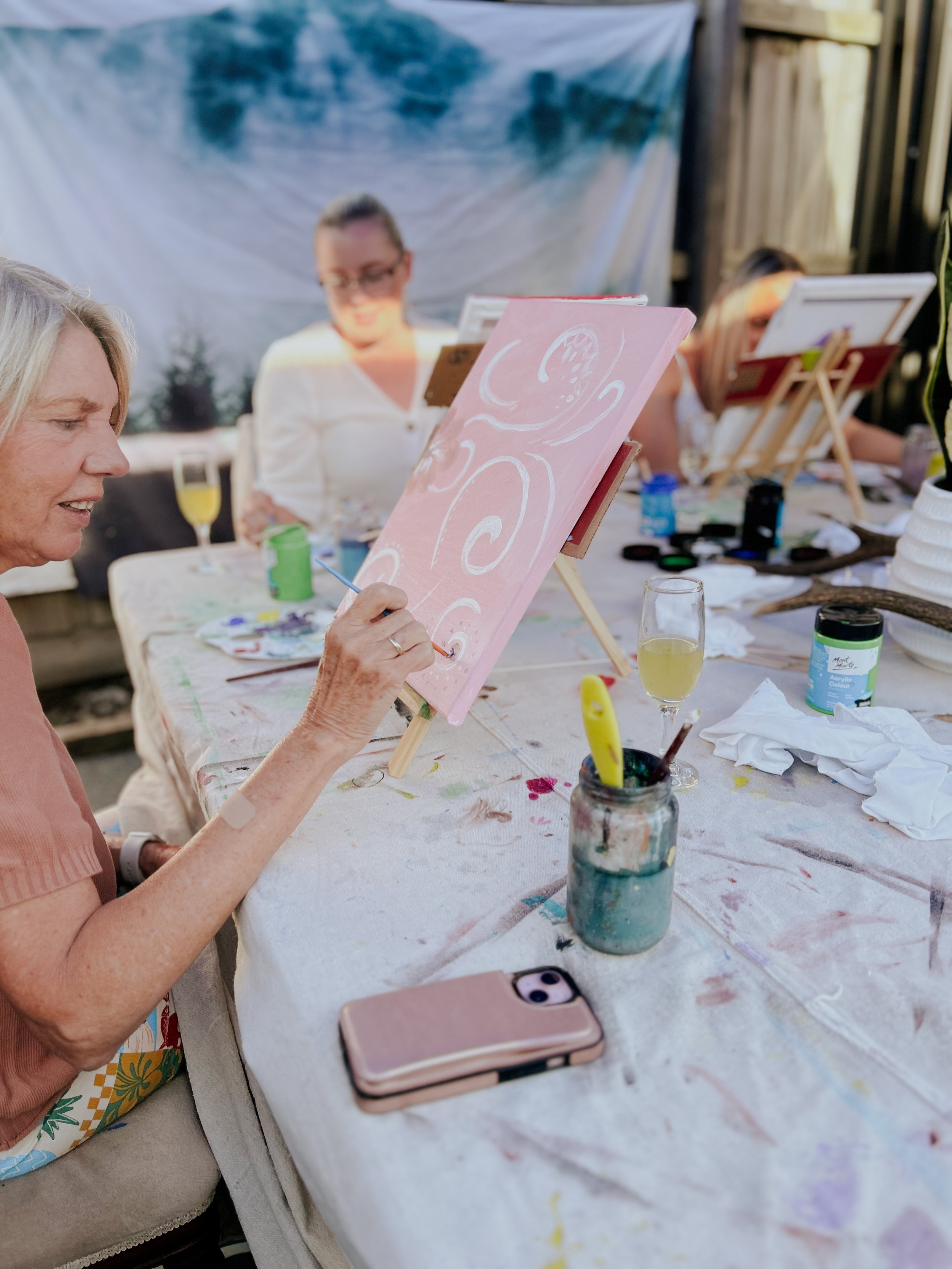 Women painting on small canvases at a table outdoors during daytime, with art supplies and drinks on the table.