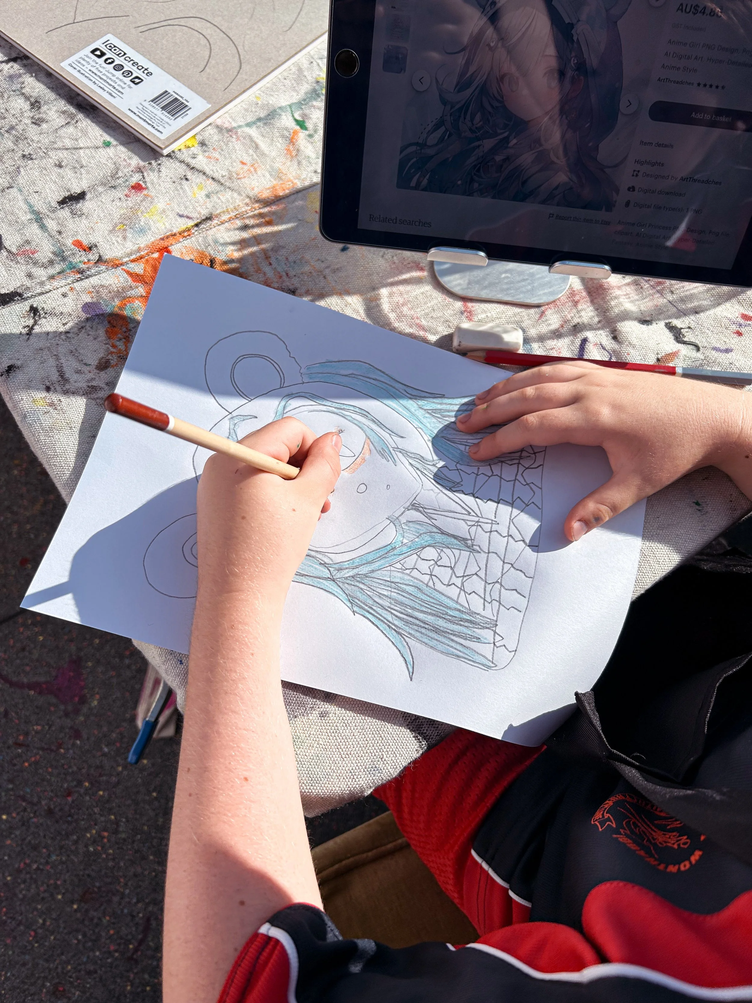 A person is drawing a colorful picture of a dragon on a piece of paper, with a computer screen displaying an anime character in the background. The person is using colored pencils and sitting at a table covered with paint splatters.