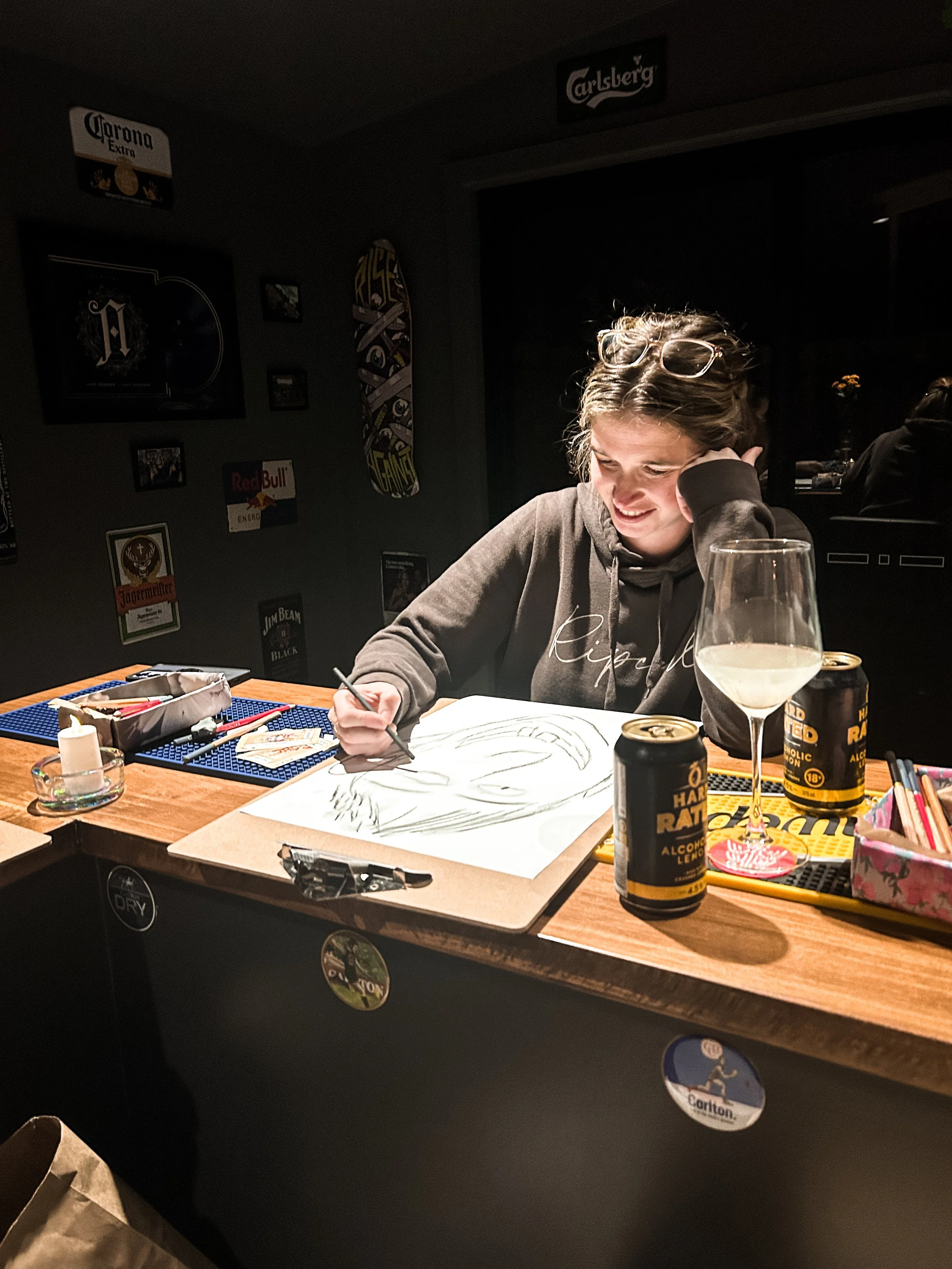 A woman drawing on paper at a table in a dimly lit room decorated with beer and liquor advertisements, with a glass of white wine and beverage cans on the table.