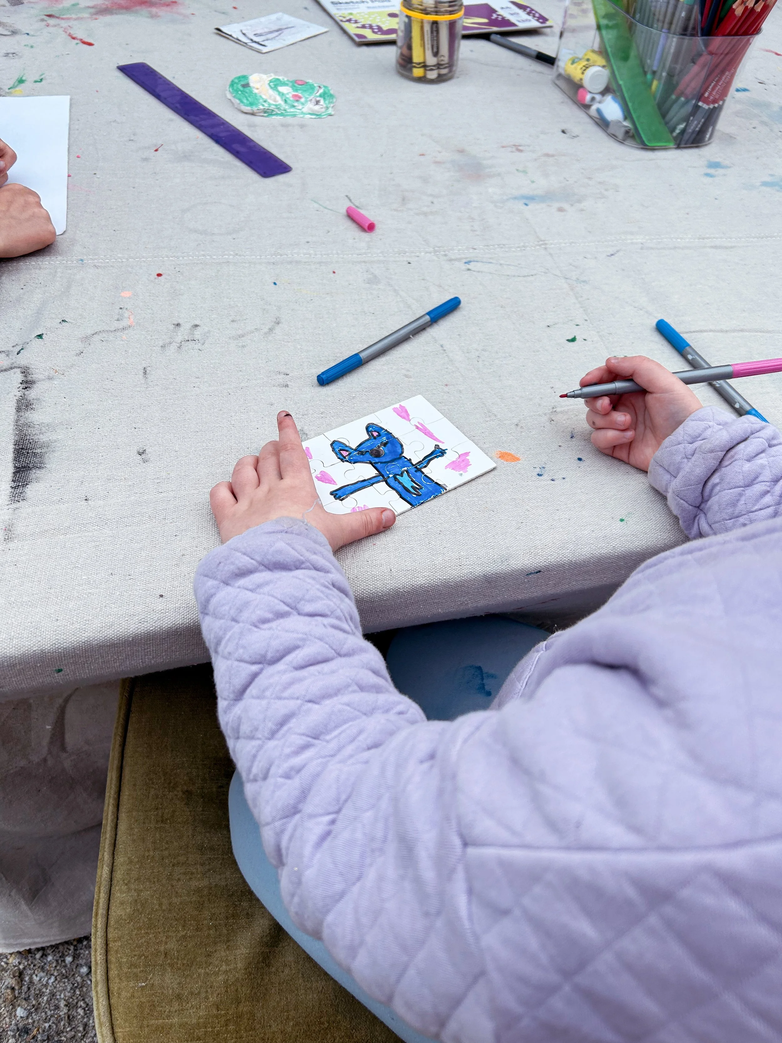 Child creating a colorful picture of an animal on a small paper with markers, seated at a table with art supplies.