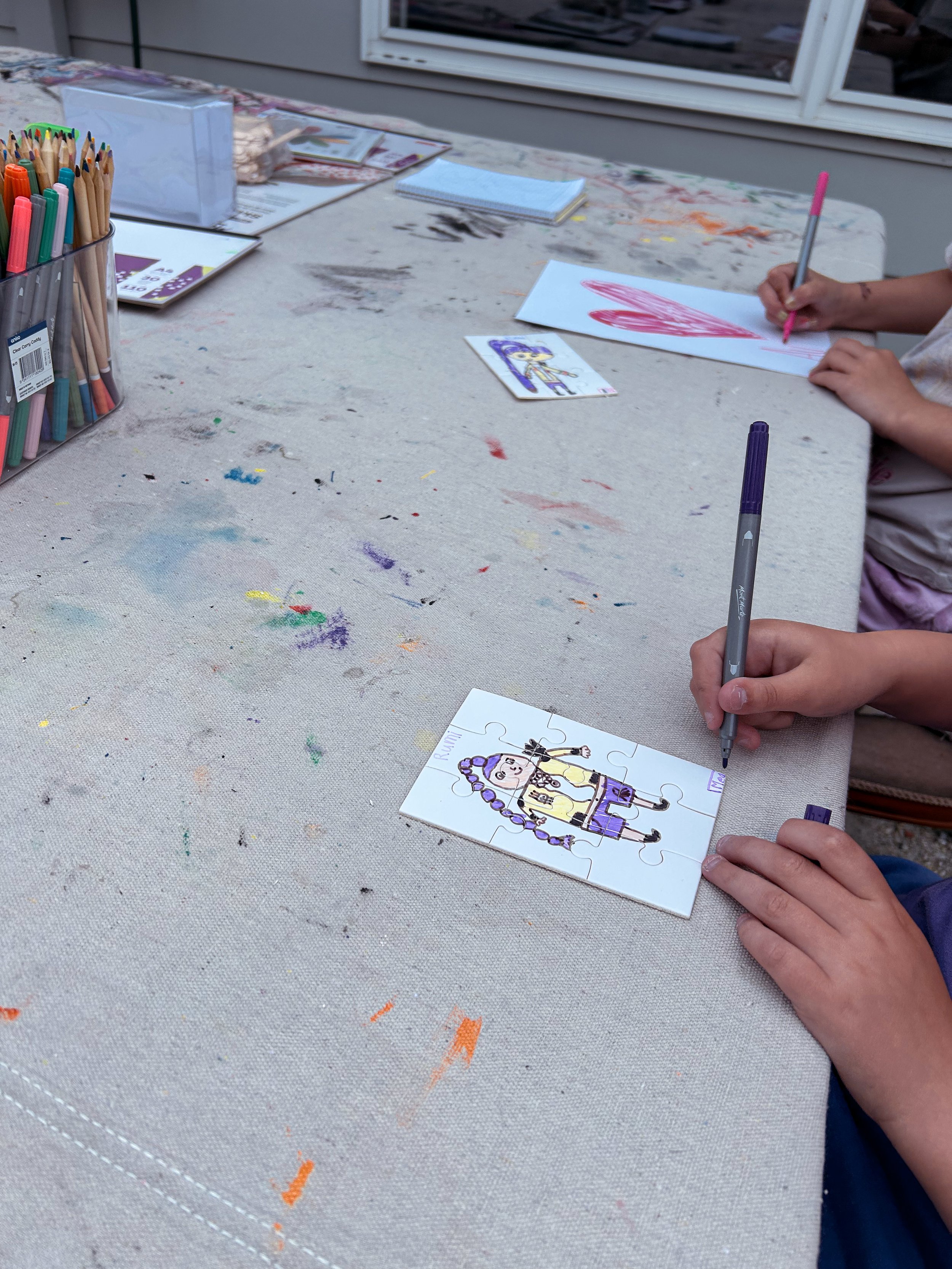 Two children are coloring on paper puzzles on a table with art supplies. One child is drawing a red heart, while the other is coloring a girl with purple hair and a yellow dress.