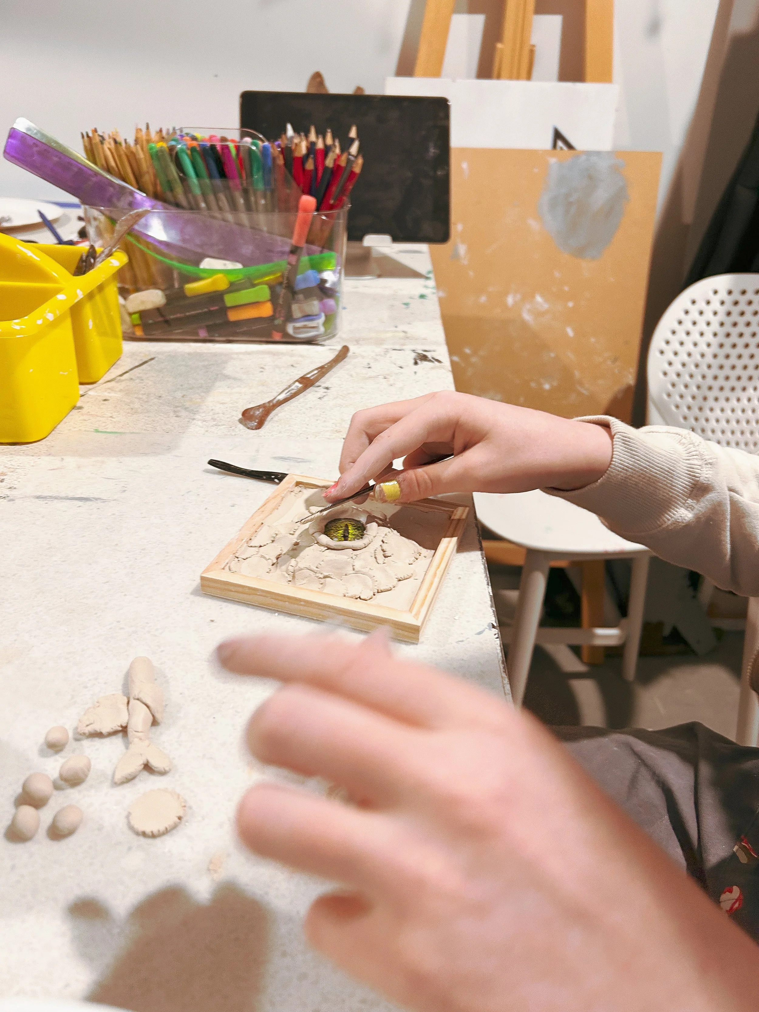 Close-up of a person working on a small wooden box with eye detail inside, surrounded by clay and ceramic pieces on a white table, with art supplies in the background.