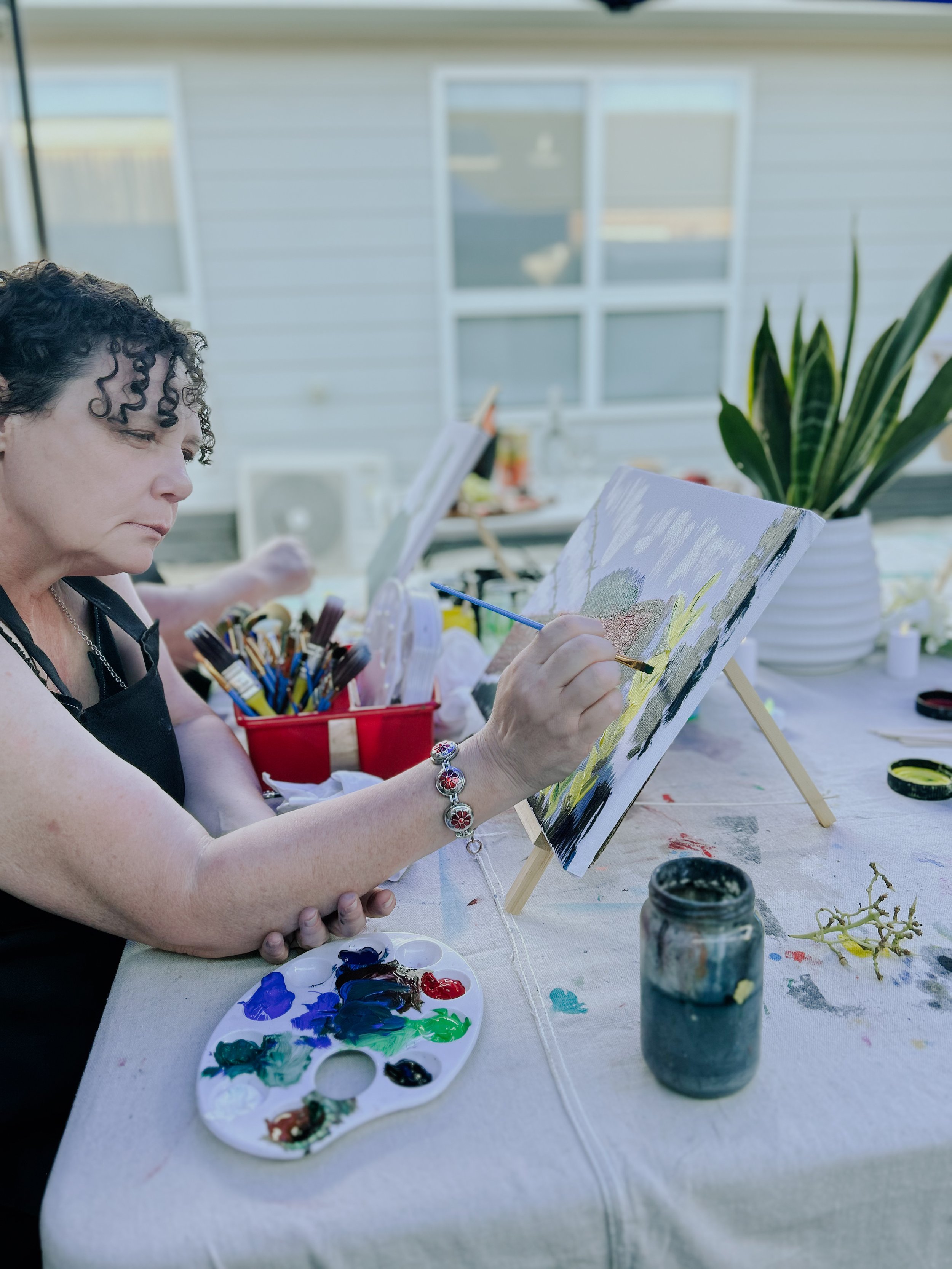 Woman painting on a canvas set on a small easel outside, with art supplies and a potted plant nearby.